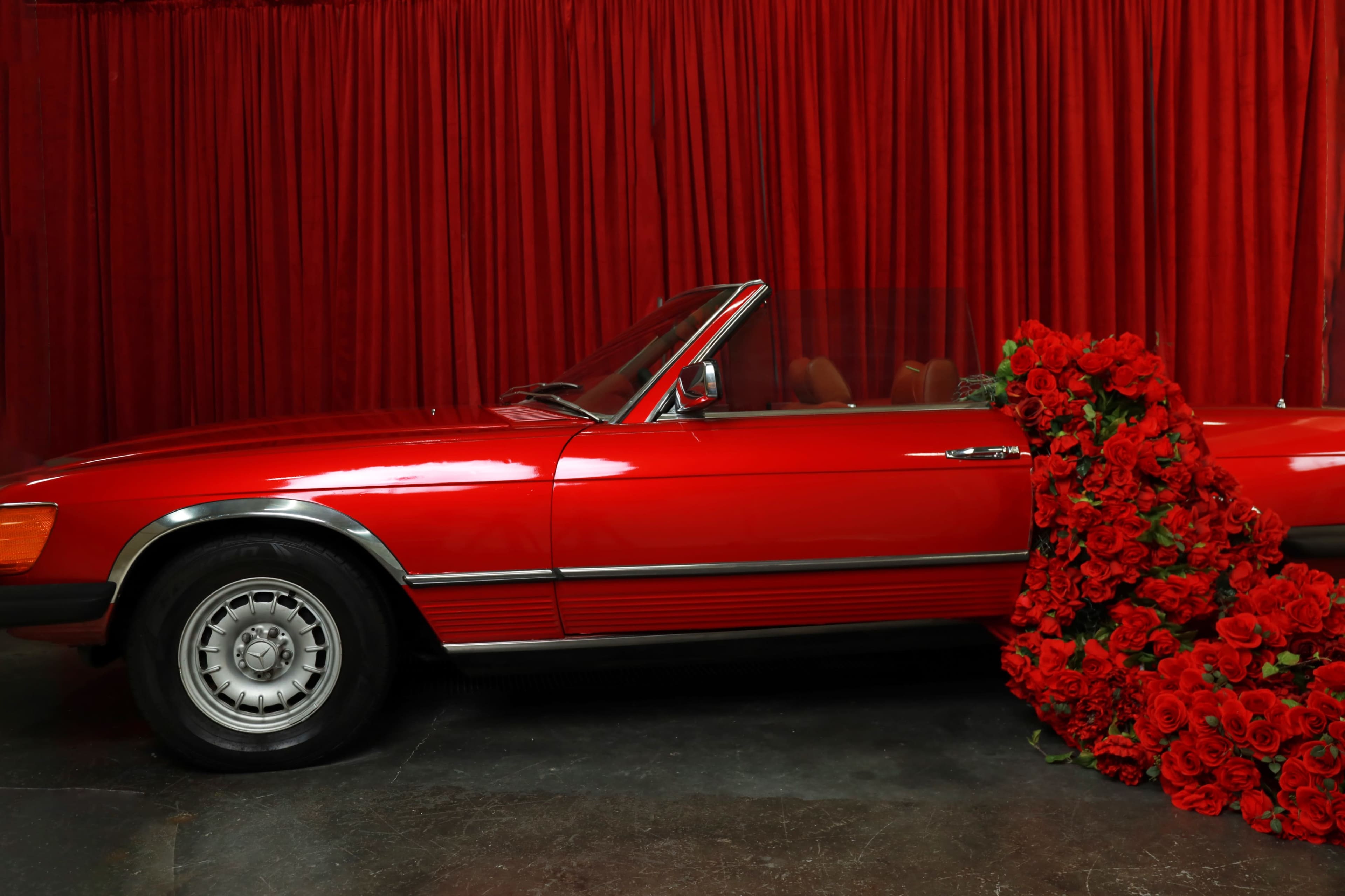 A red convertible car is partially covered by a large cluster of red roses against a backdrop of red curtains.