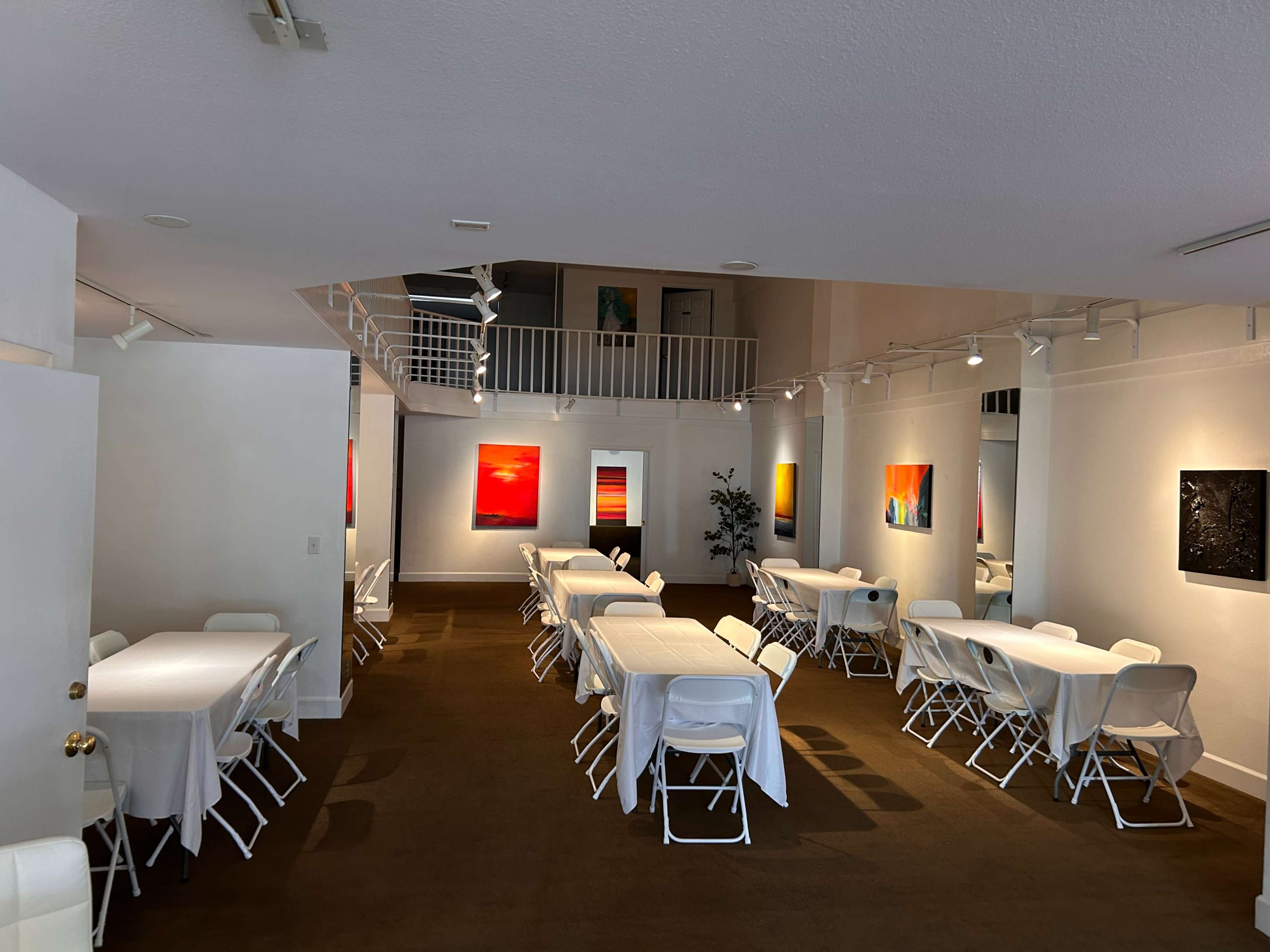 The image shows an interior space set up for an event, featuring rows of tables covered with white tablecloths and walls adorned with colorful paintings.