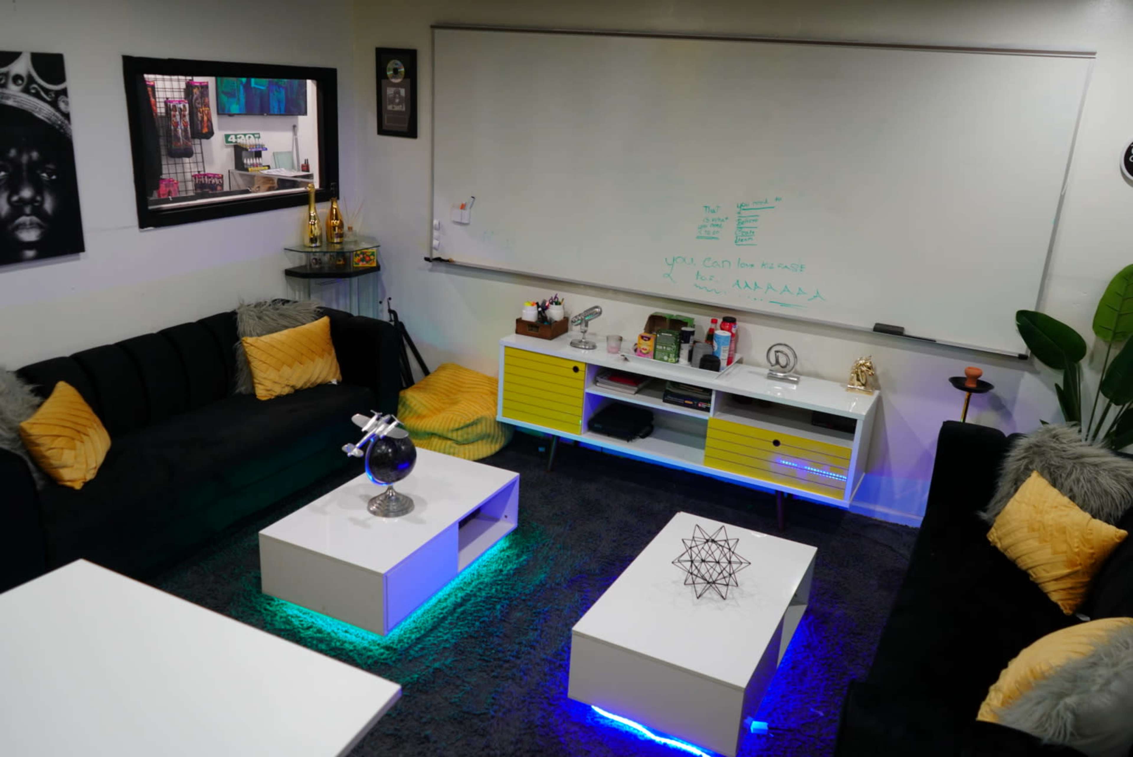 A modern lounge area with black sofas, white coffee tables featuring LED lighting, and a whiteboard mounted on the wall.