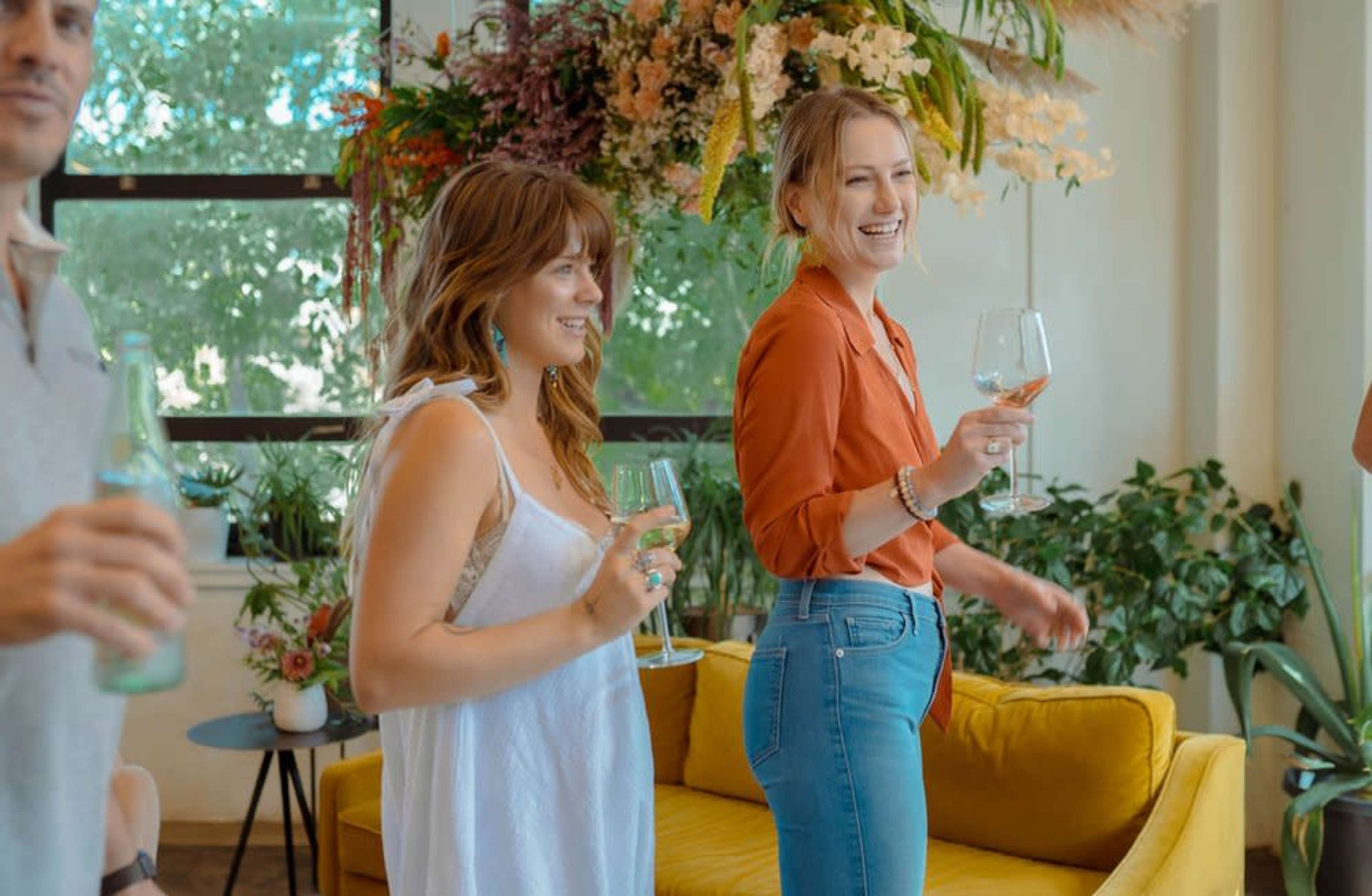 Two women are holding glasses, smiling and engaging, while standing in a brightly lit room filled with plants and a yellow sofa.