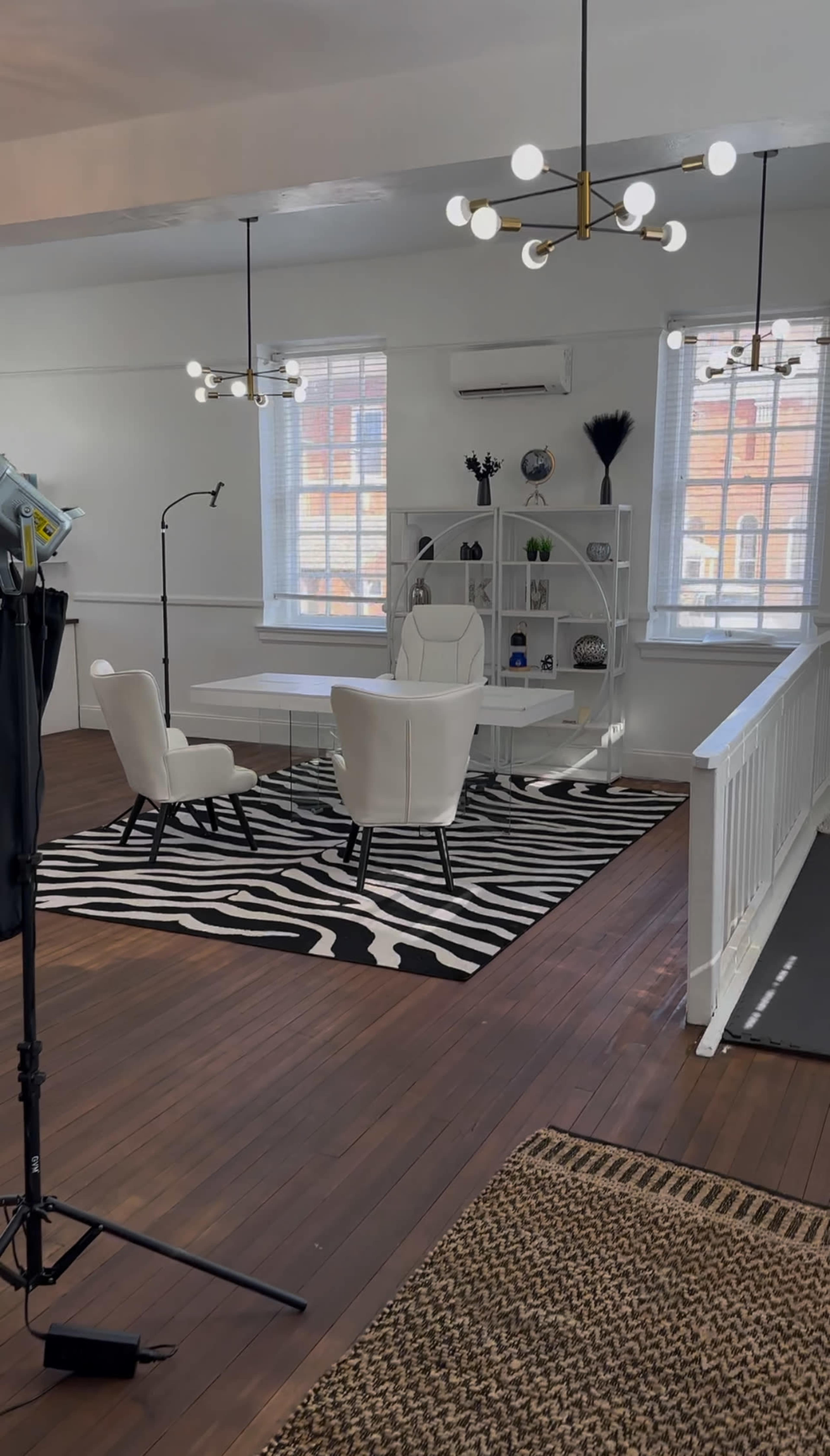 The image shows a bright, modern office space featuring a glass desk, two white chairs, a striped rug, and decorative shelves, with large windows allowing natural light to fill the room.