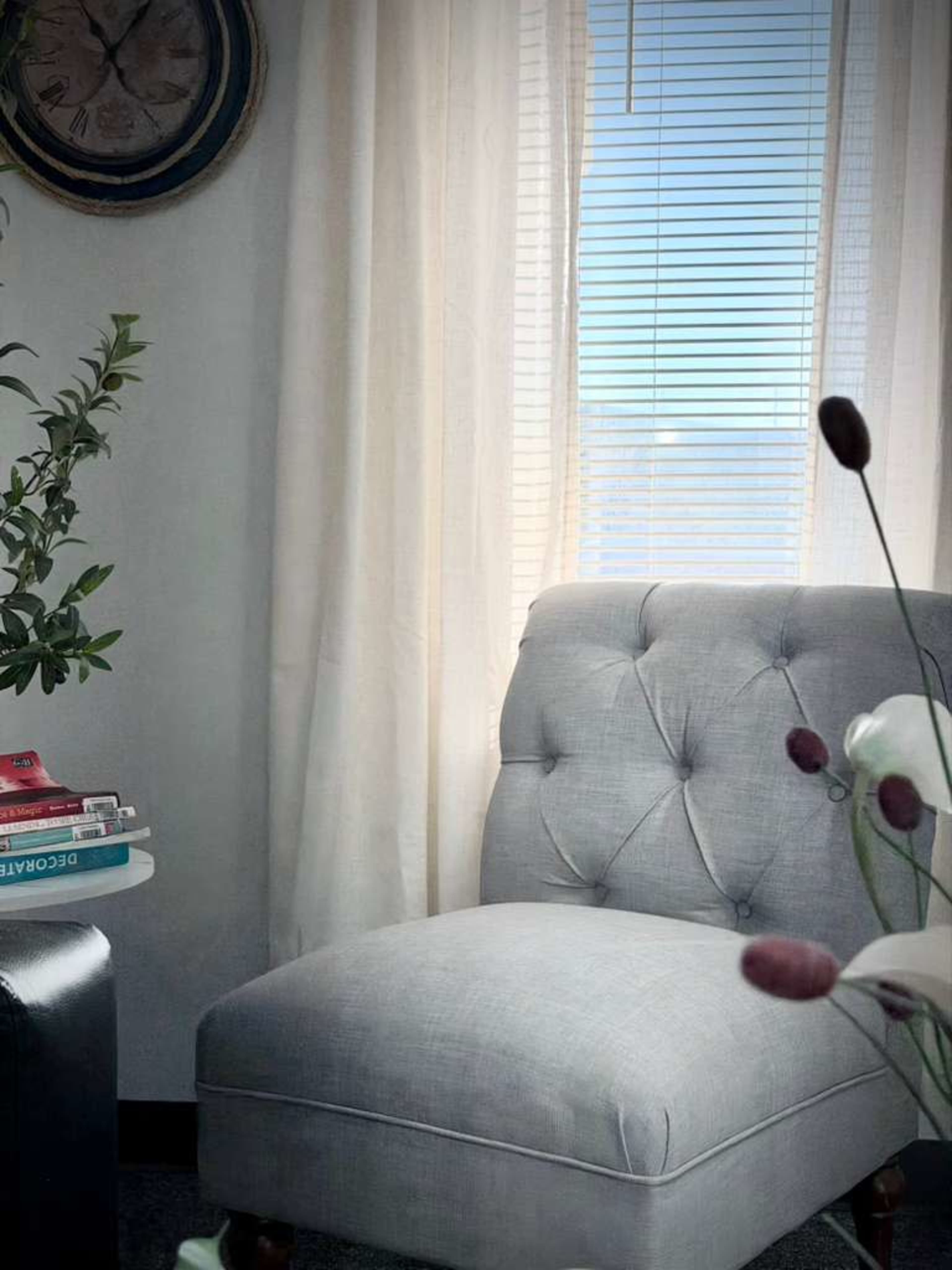 A gray upholstered armchair is positioned beside a window with blinds, near a small table with stacked books and a decorative plant.