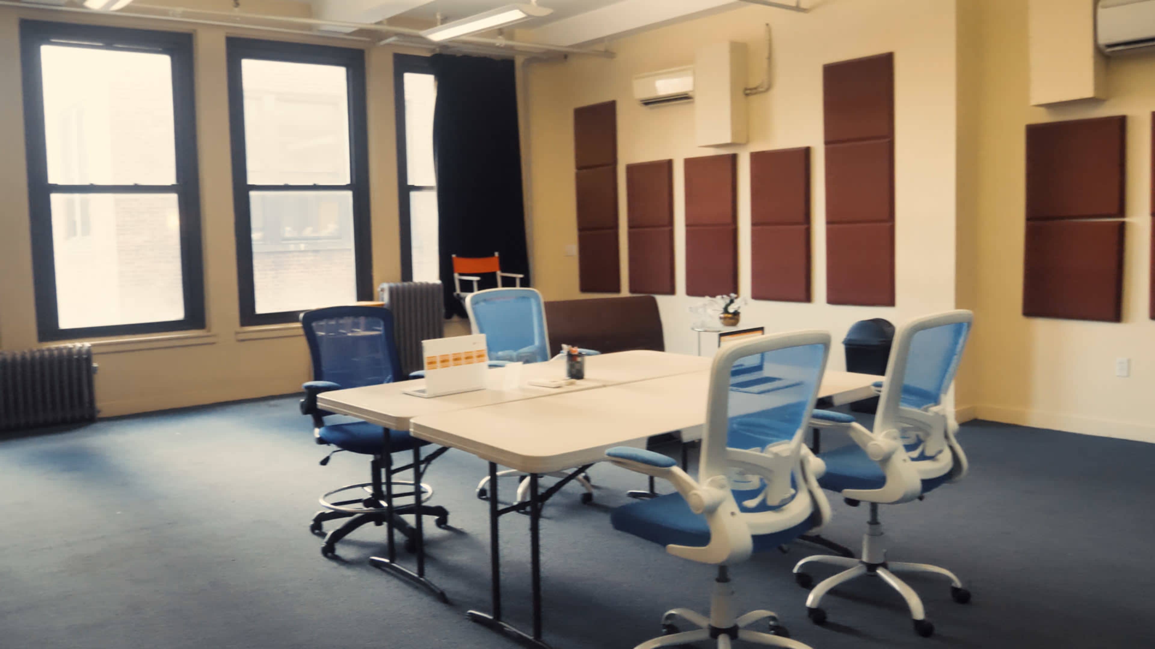 A meeting room features a rectangular table surrounded by blue office chairs, with large windows and sound panels on the walls.