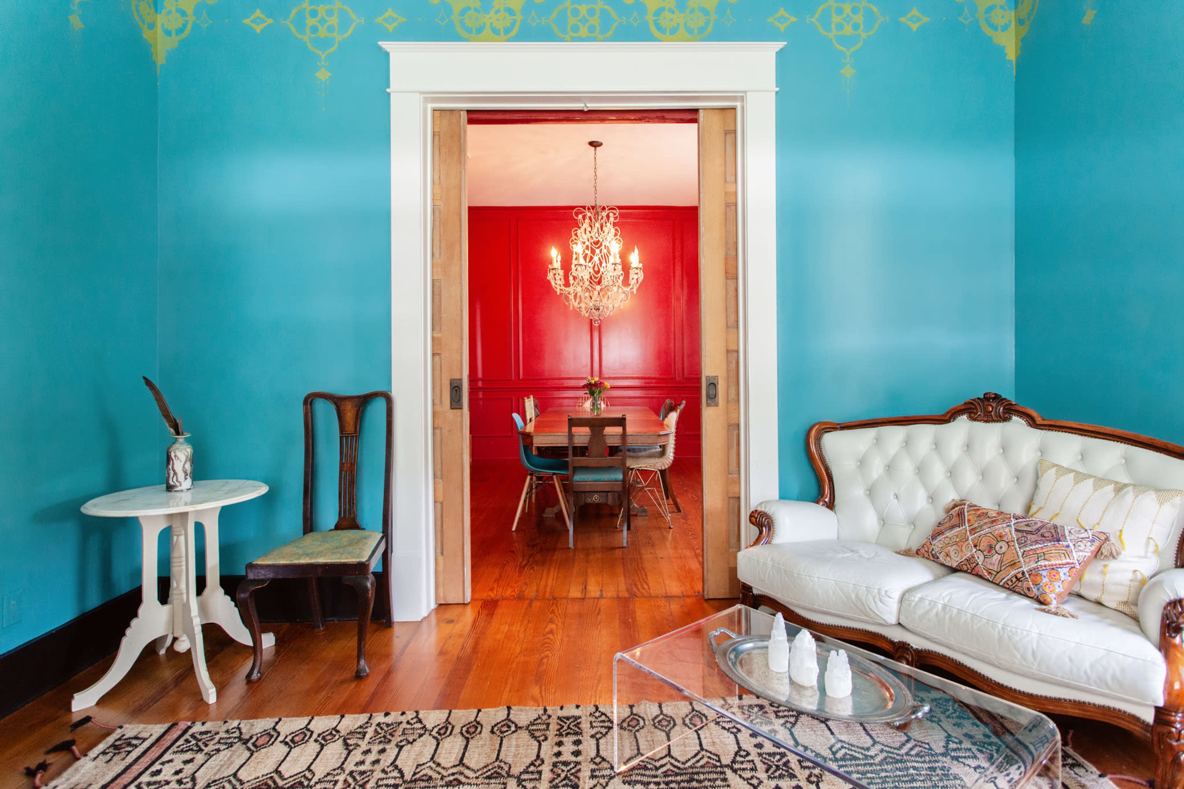 The image features a brightly colored room with a turquoise wall, showcasing a white sofa and a decorative table, while an adjacent dining area with a red wall and a chandelier is visible through an open doorway.