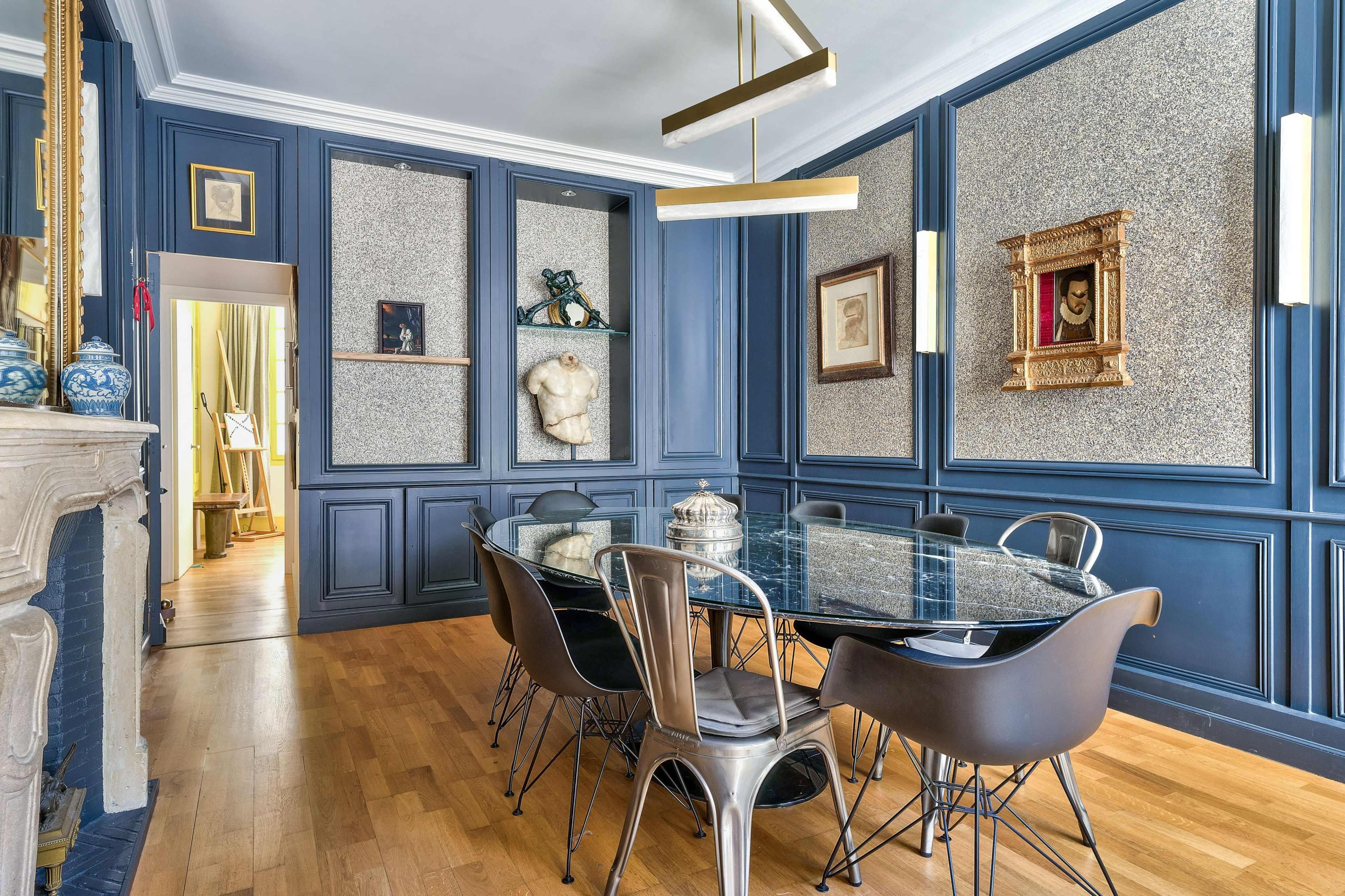 The image shows a dining room with a glass table surrounded by black chairs, blue paneled walls, and decorative artwork displayed on the walls.