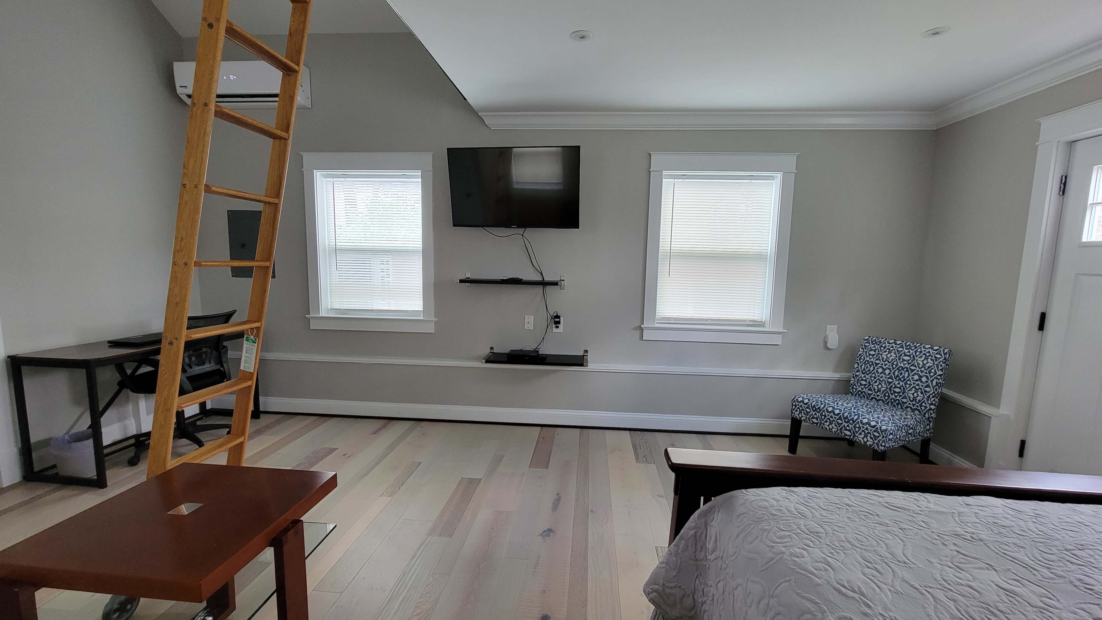 The image shows a modern bedroom with a wooden ladder, a wall-mounted TV, a small table, a blue patterned chair, and two windows.