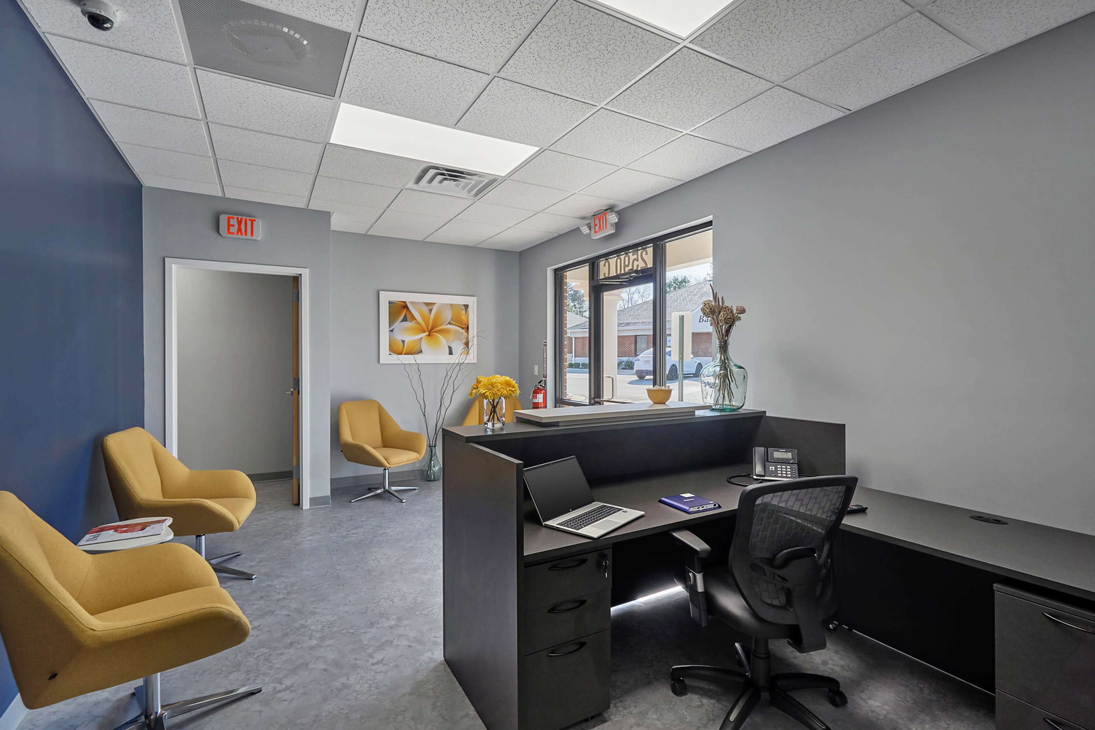 A modern office reception area featuring two yellow chairs, a black desk with a chair, and a large window letting in natural light.