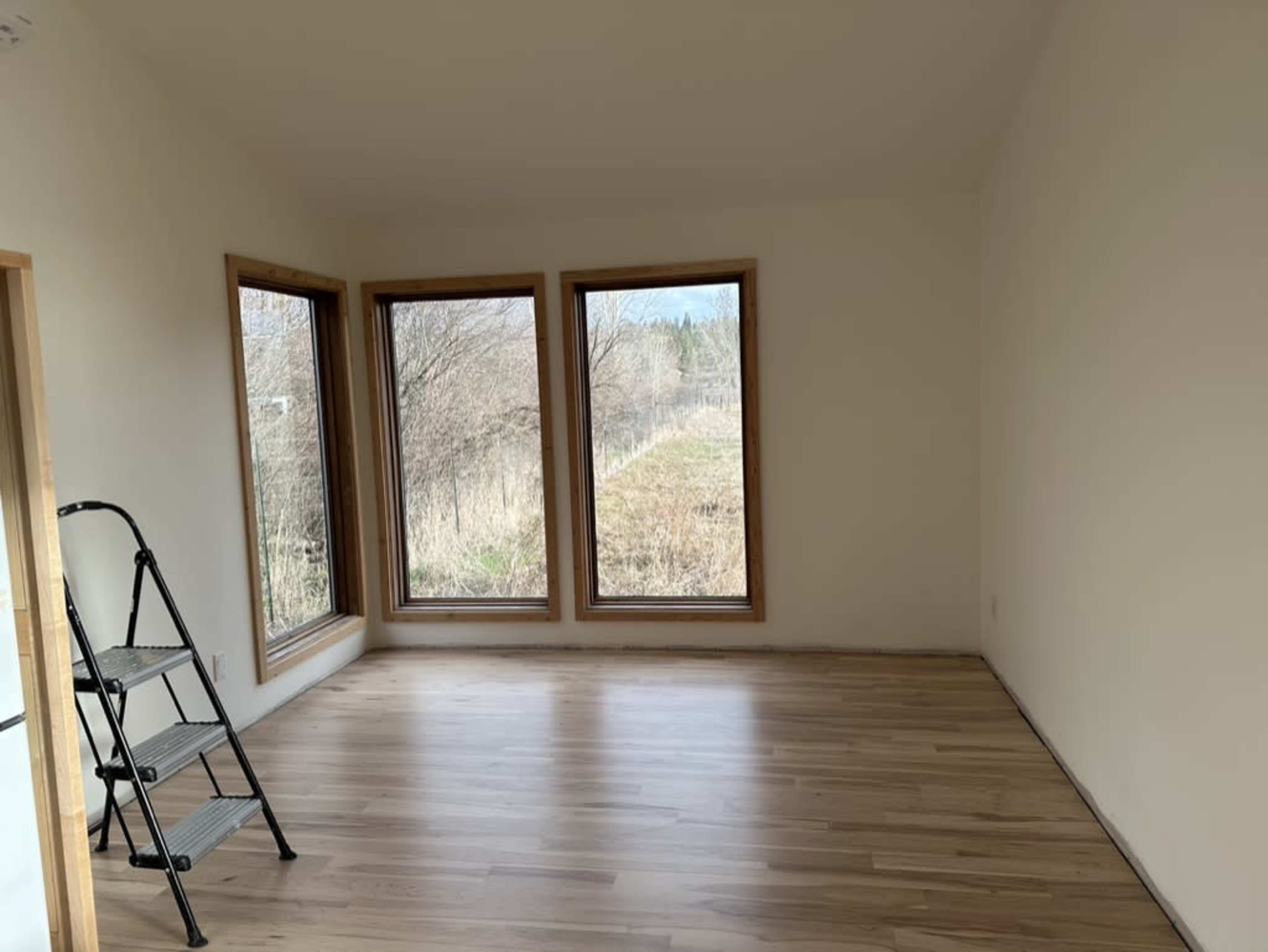 A brightly lit room features large windows overlooking a grassy area, with a step ladder positioned against one wall.