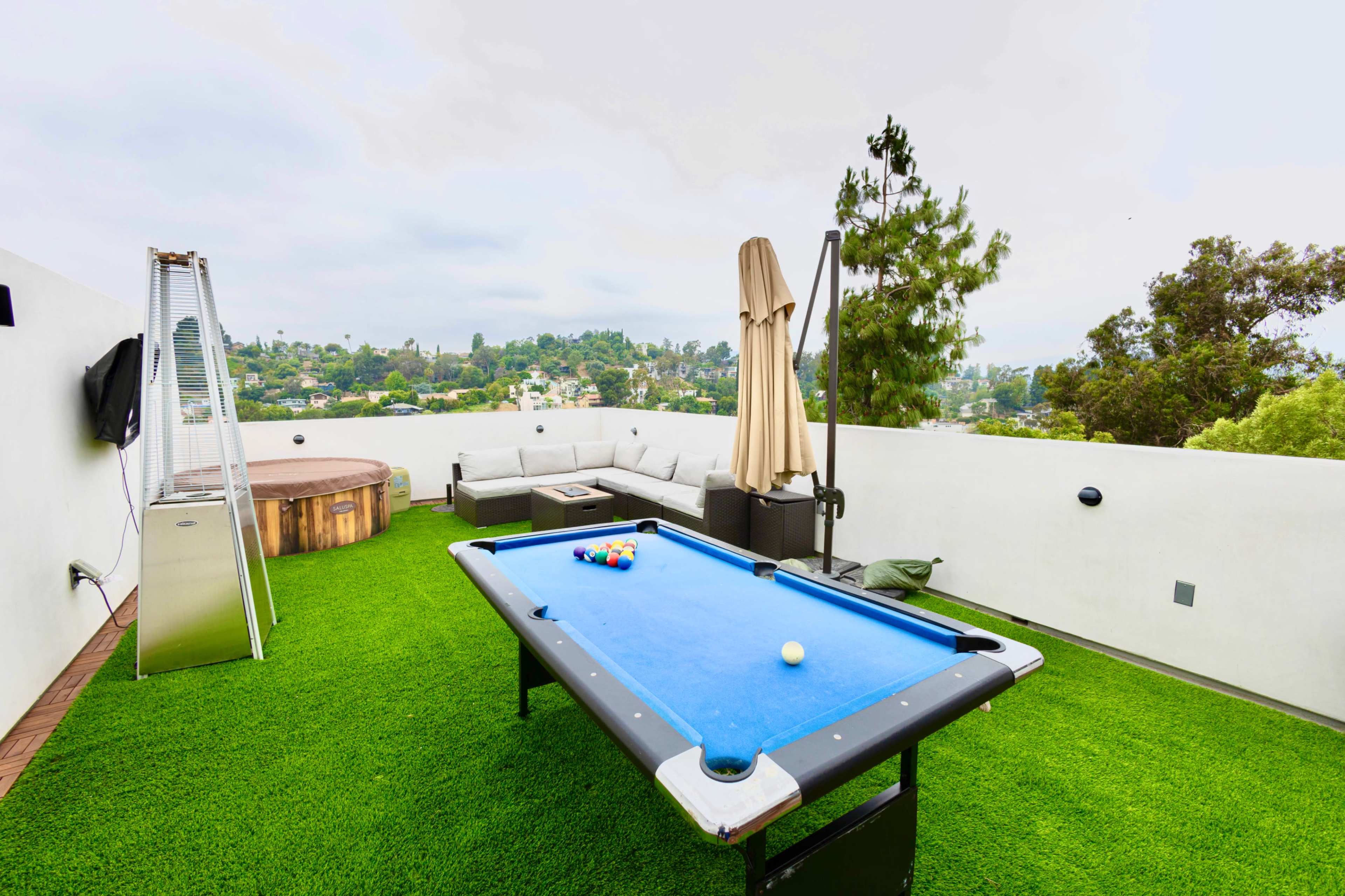 A rooftop patio features a blue pool table, a hot tub, a shaded seating area, and a view of the surrounding landscape.