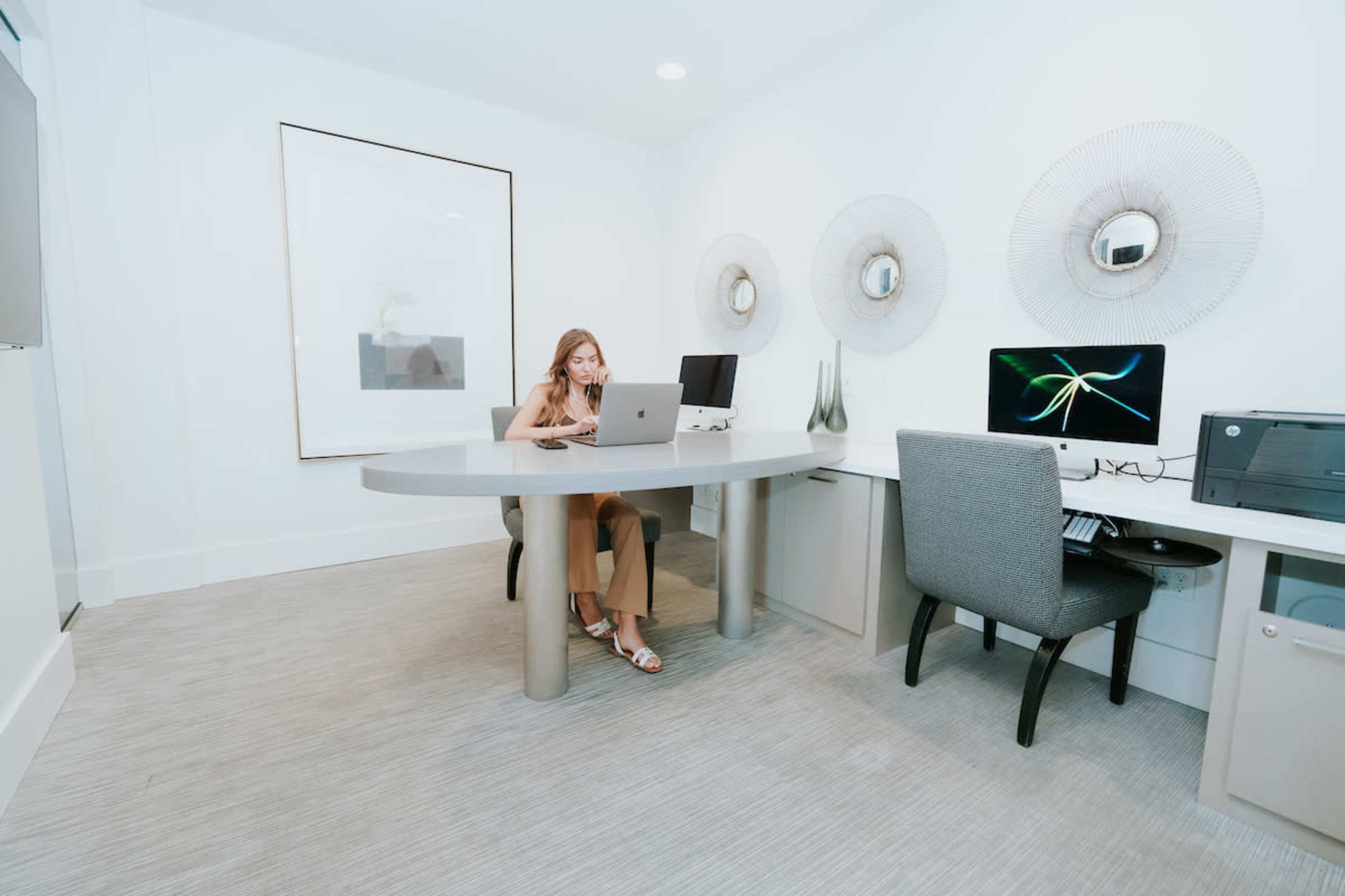 A woman sits at a round desk in a modern office space, working on a laptop, with two computers and decorative wall elements in the background.