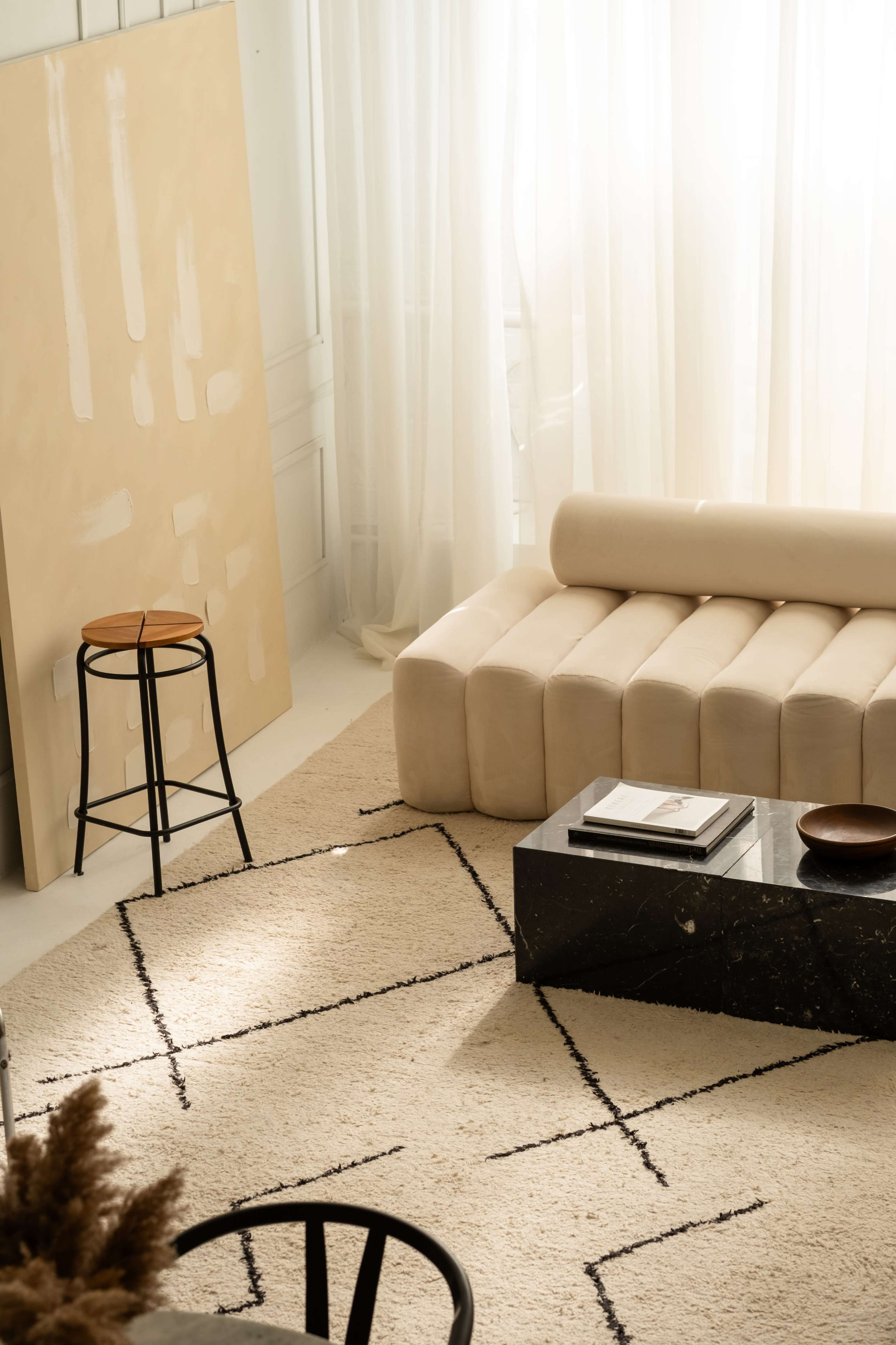 The image shows a modern living room featuring a beige curved sofa, a black coffee table, a neutral rug with geometric patterns, and large windows draped with sheer curtains.