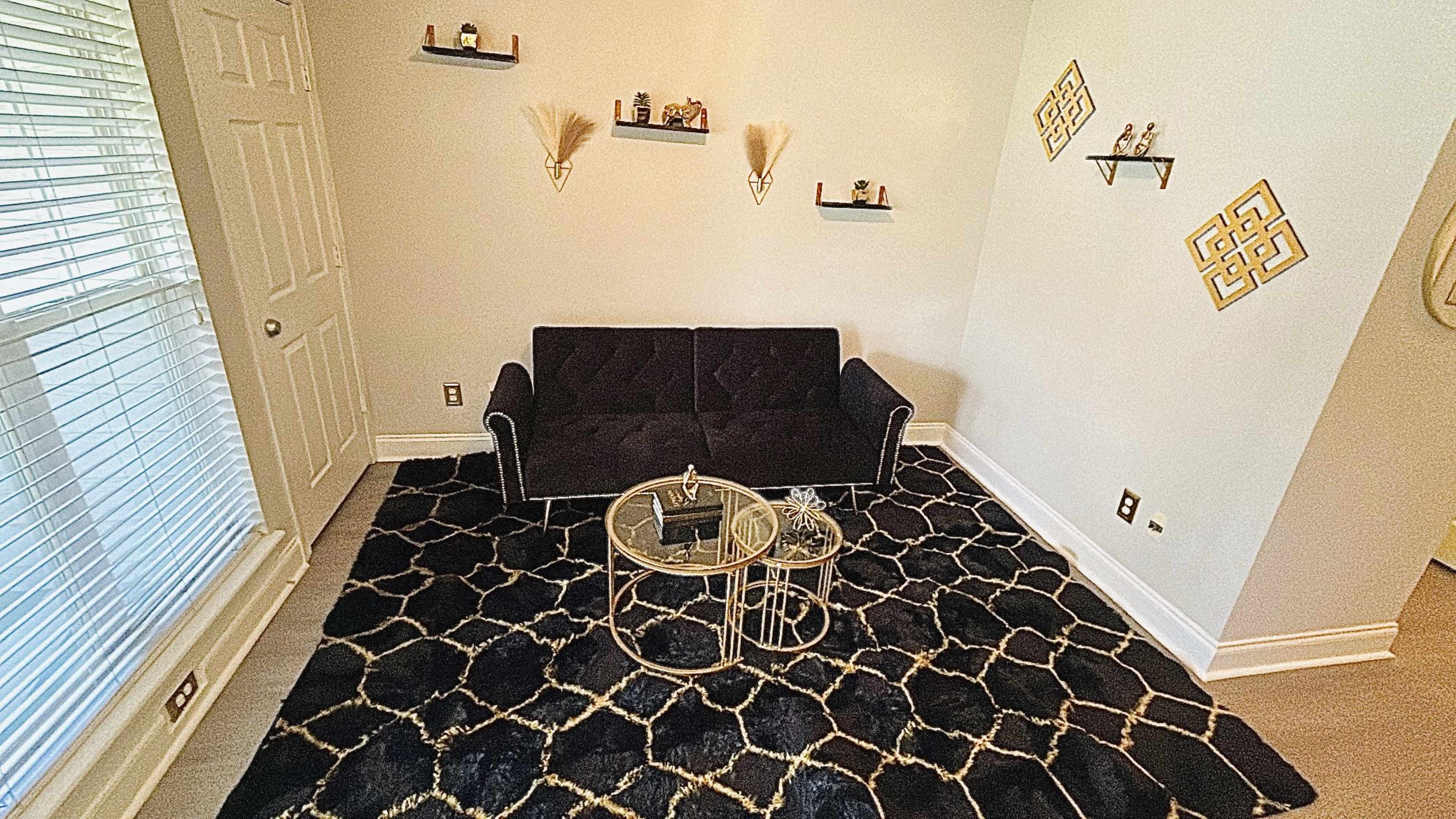 The image shows a living room with a black sofa, a circular glass coffee table, and decorative wall shelves.