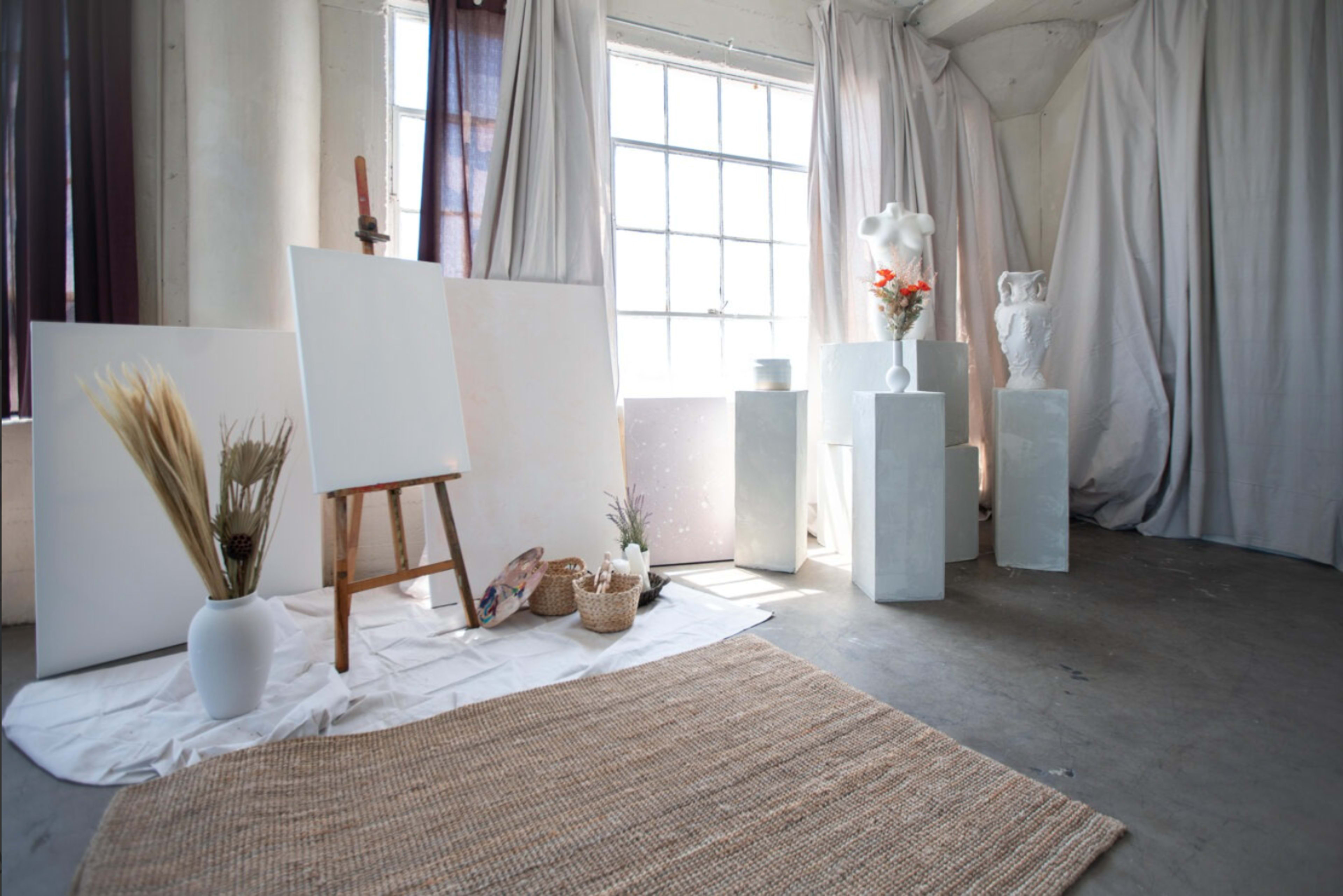 A bright art studio with an easel, blank canvases, decorative pedestals, and a woven rug on a concrete floor.