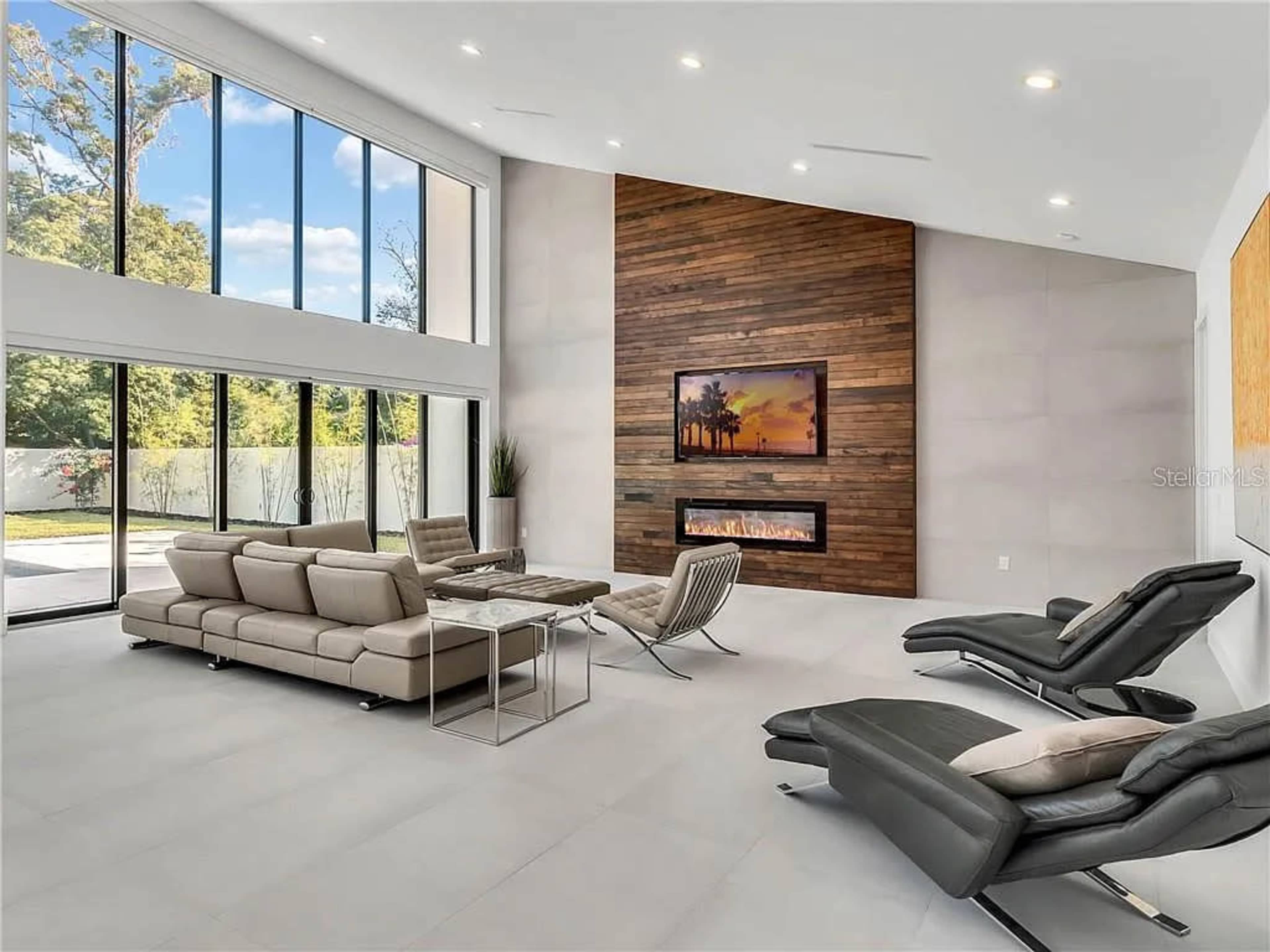 The modern living room features large windows, a wood-paneled accent wall with a fireplace, and sleek furniture arranged for relaxation.