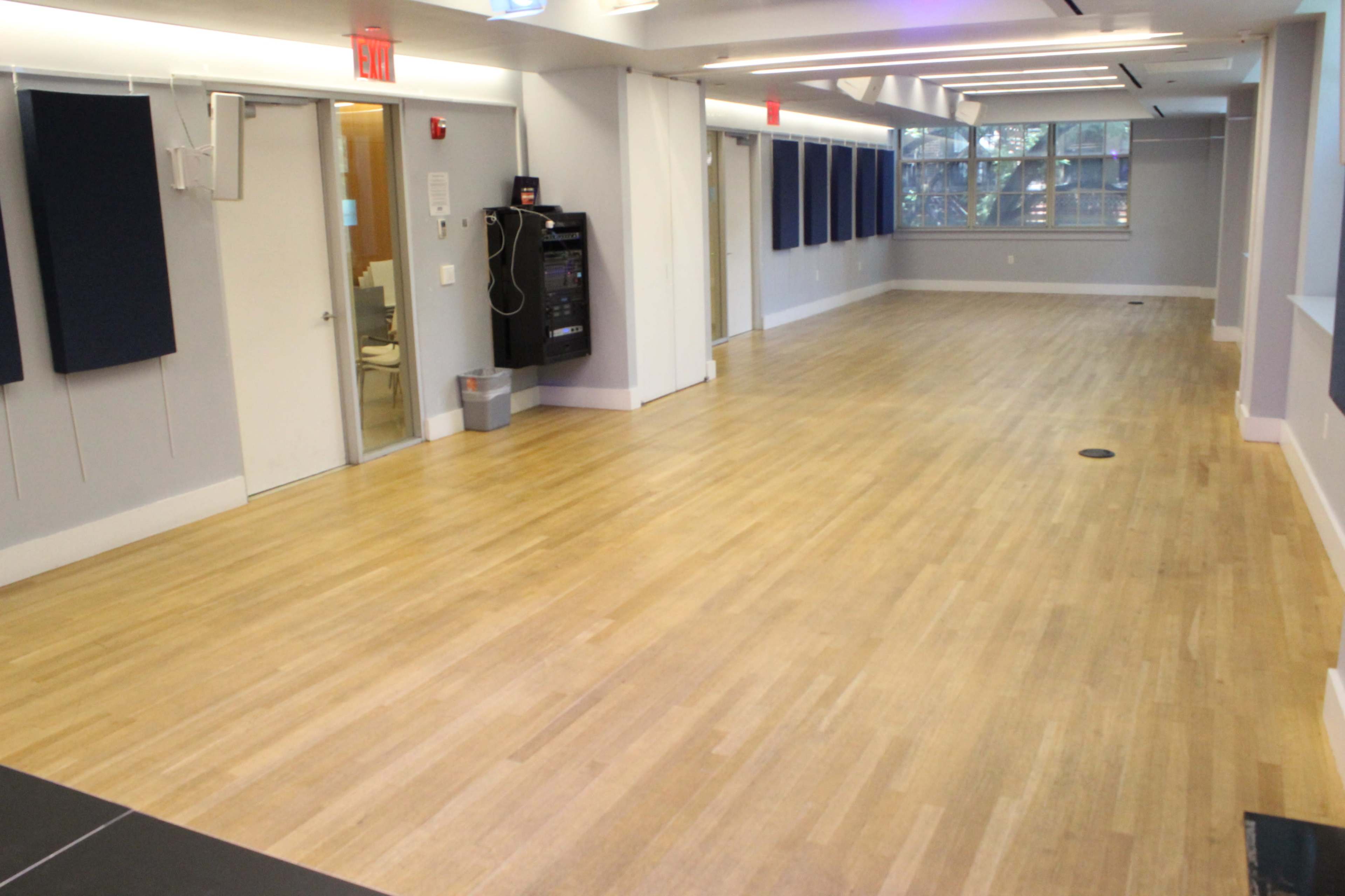 The image shows a spacious, empty room with wooden flooring, large windows, and several doors along the walls.