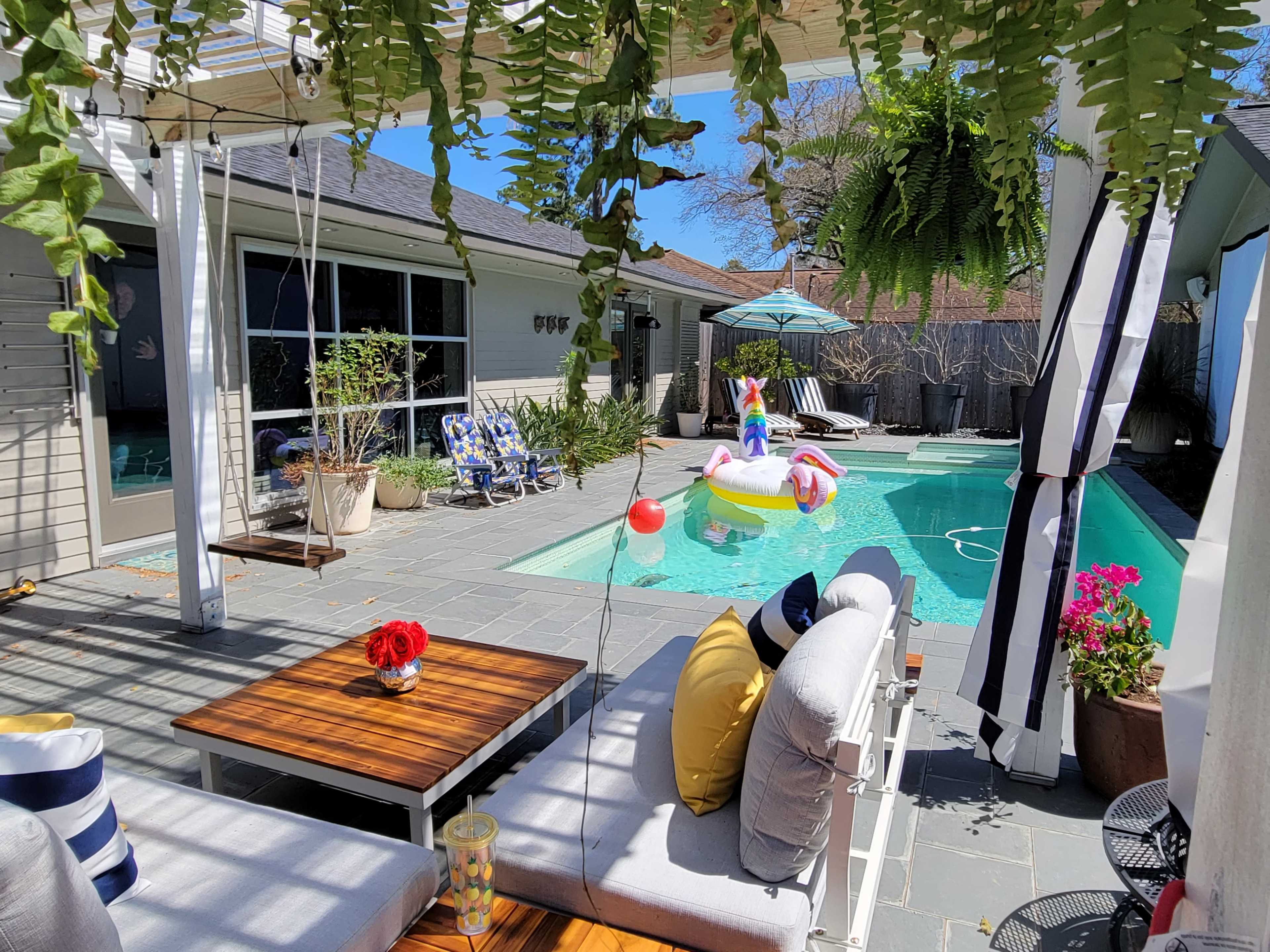 A sunny backyard features a swimming pool surrounded by lounge chairs, potted plants, and a child playing on a float.