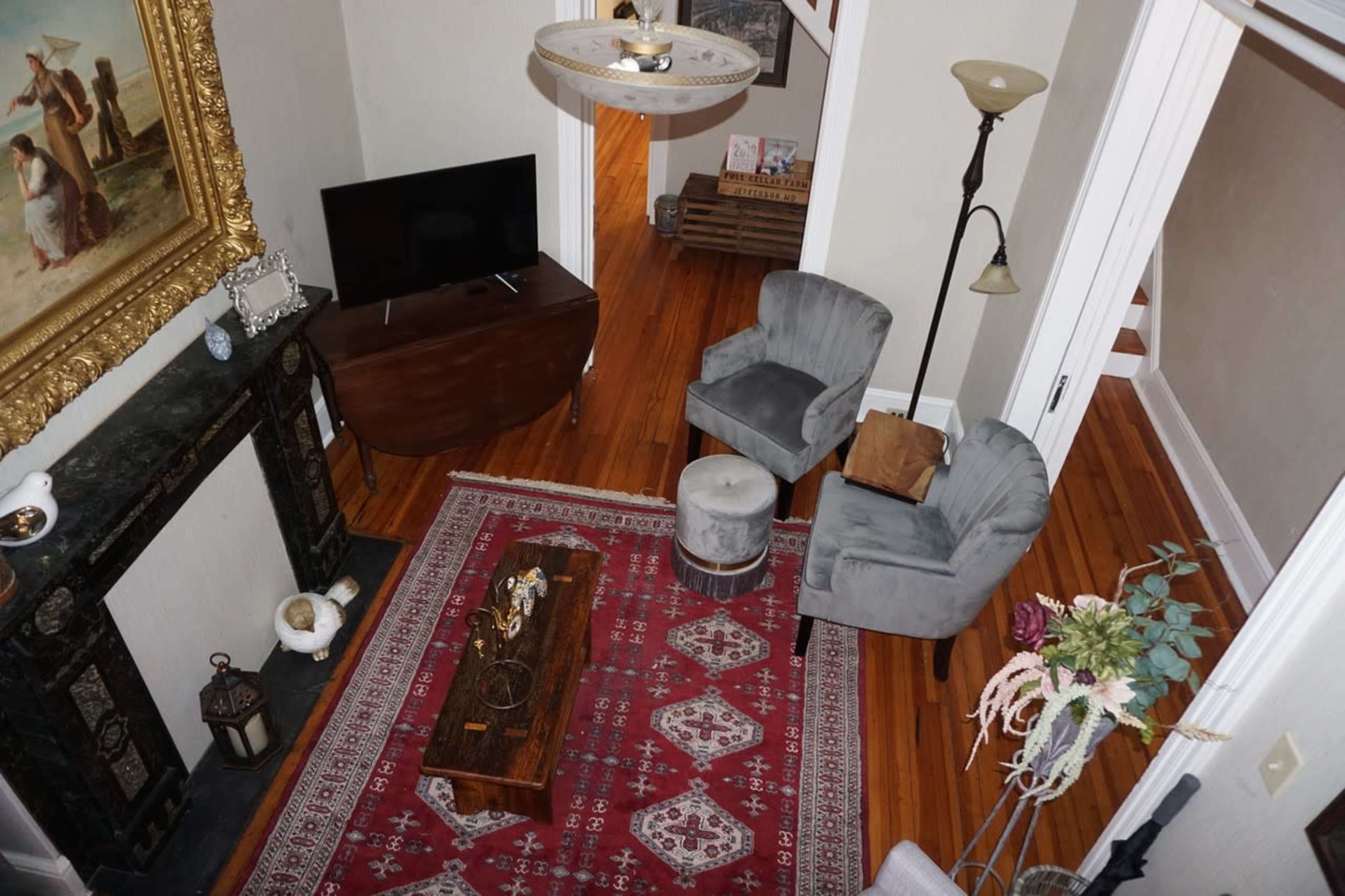 A living room with a television, a wooden coffee table, two gray chairs, and a large painting above a fireplace, all arranged on a red area rug.