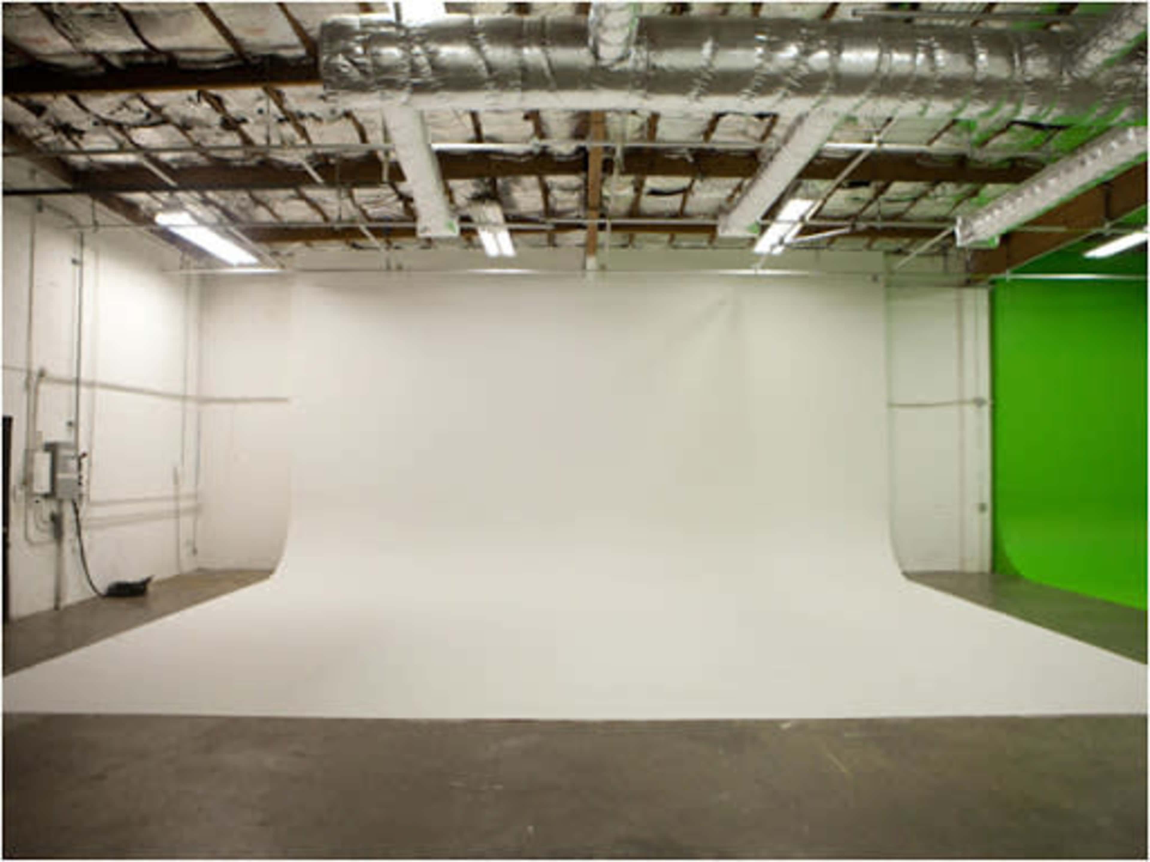 A spacious studio with a seamless white backdrop and a green screen on opposite walls, surrounded by exposed ceiling and ductwork.
