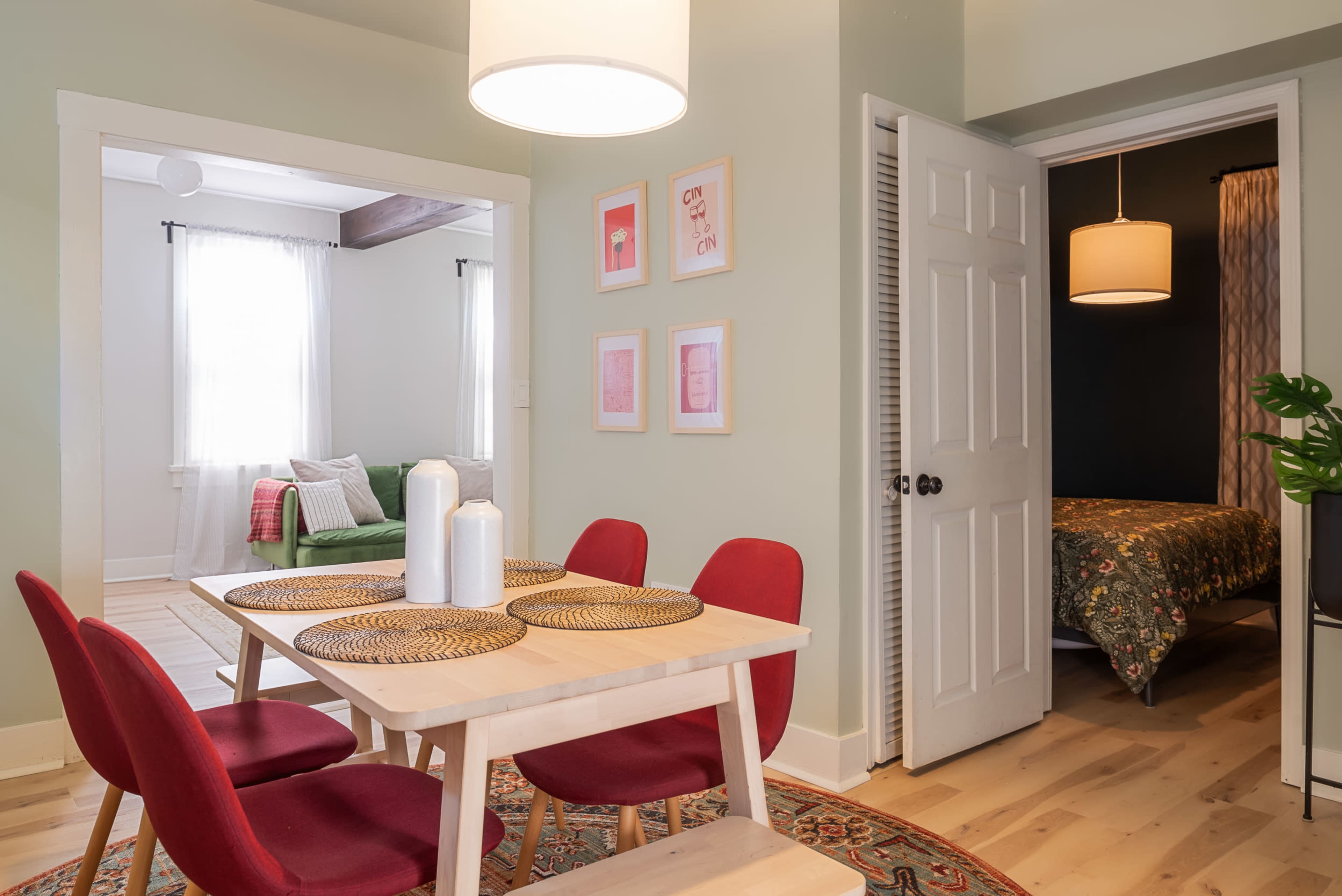 A dining area with a wooden table and red chairs is seen adjacent to a room with a bed and a green sofa in the background.