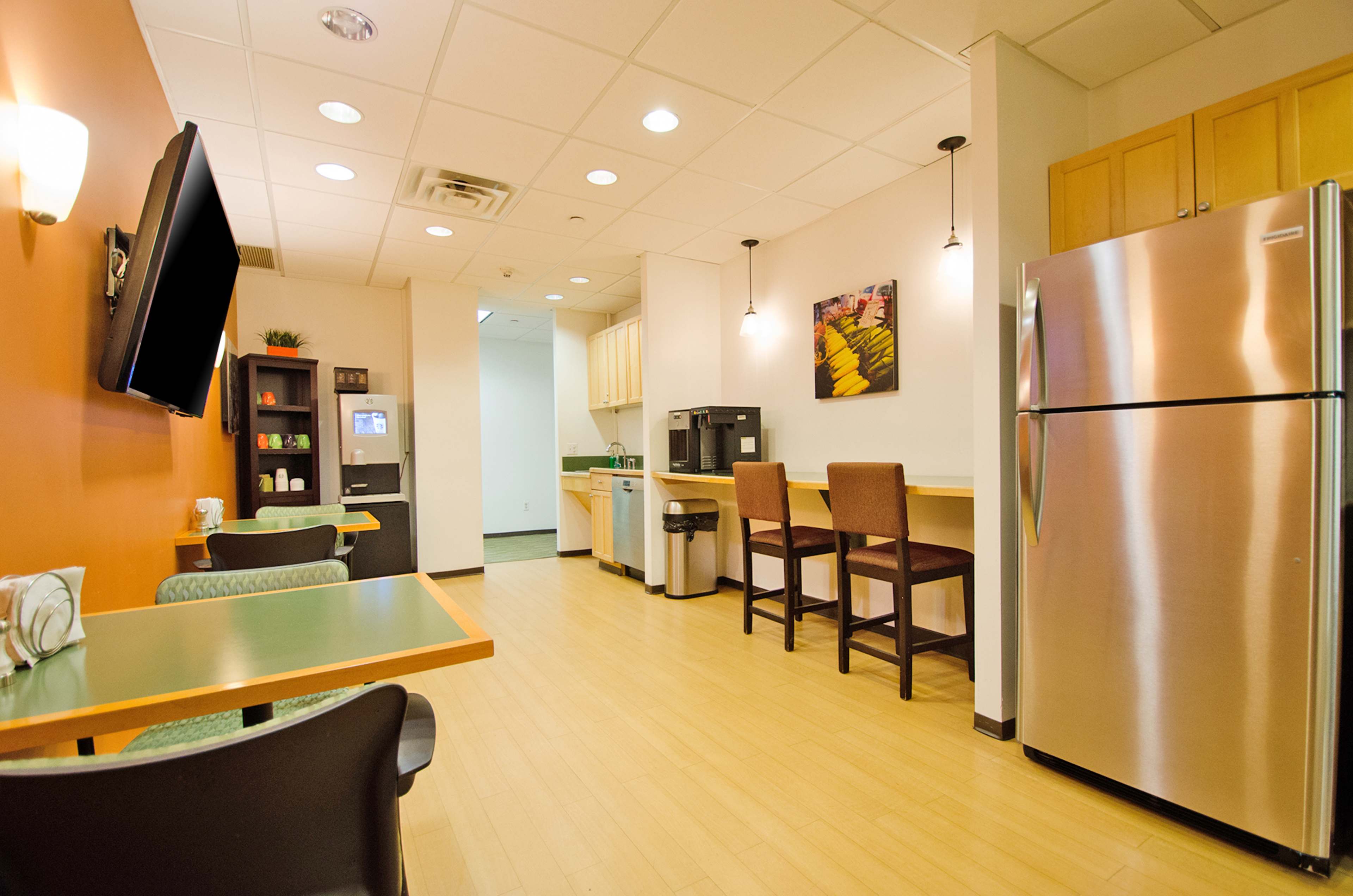 A modern kitchenette area with a refrigerator, coffee maker, seating options, and flat-screen TV along the wall.