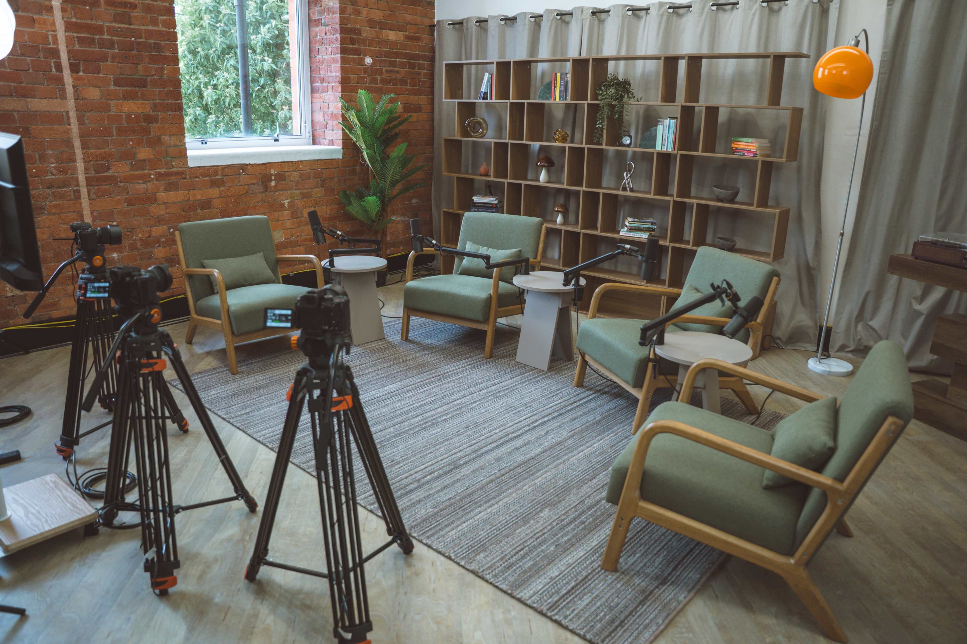 A modern studio setup features four tripods with cameras positioned around a circular seating arrangement of green chairs and small tables against a backdrop of a brick wall and a window.
