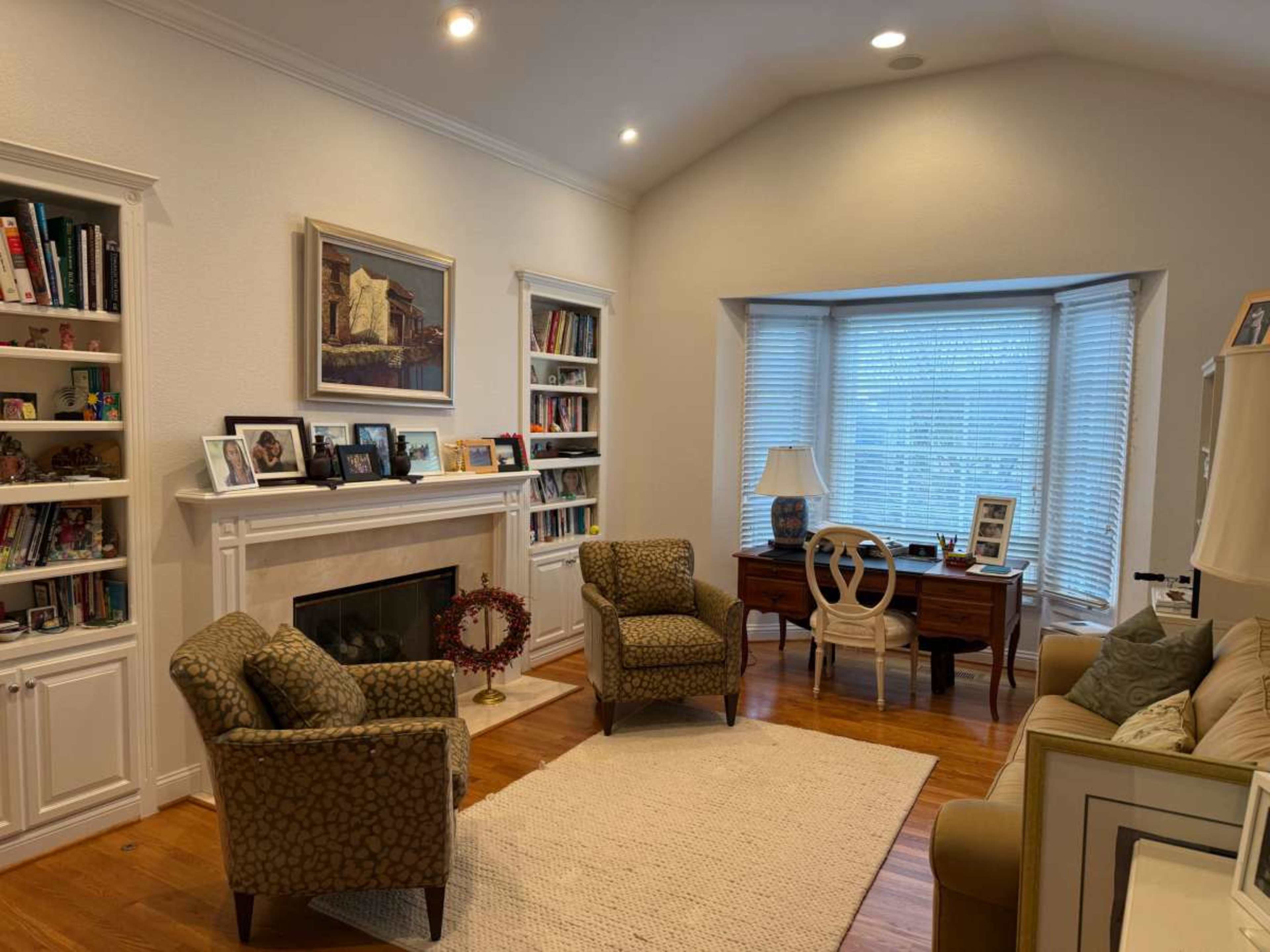 A living room features two patterned chairs, a wooden desk with a chair, a fireplace with a mantel displaying framed photos, and built-in bookshelves.