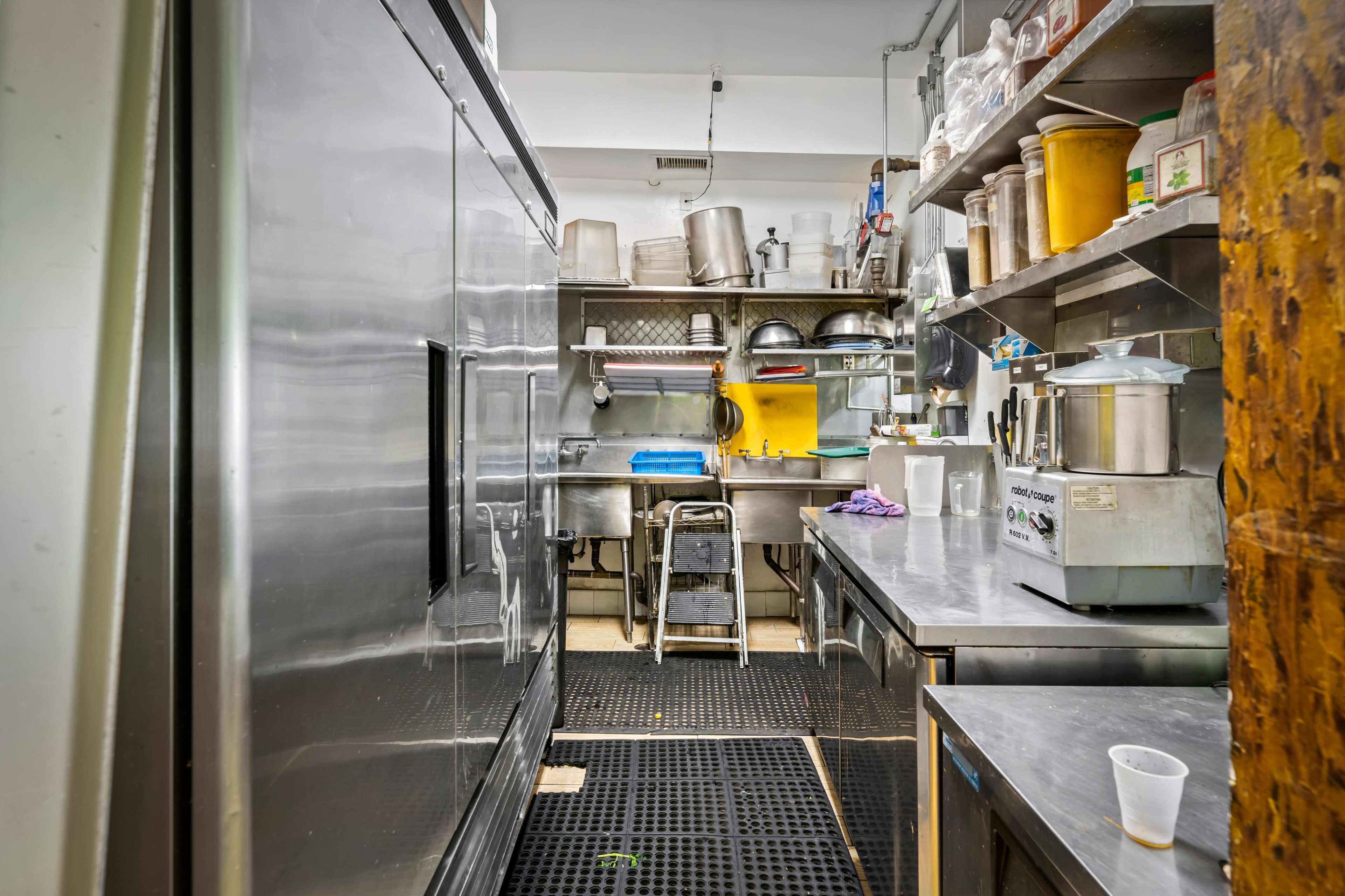 A narrow kitchen with stainless steel equipment, shelves filled with utensils and containers, and a side entrance to the cooking area.
