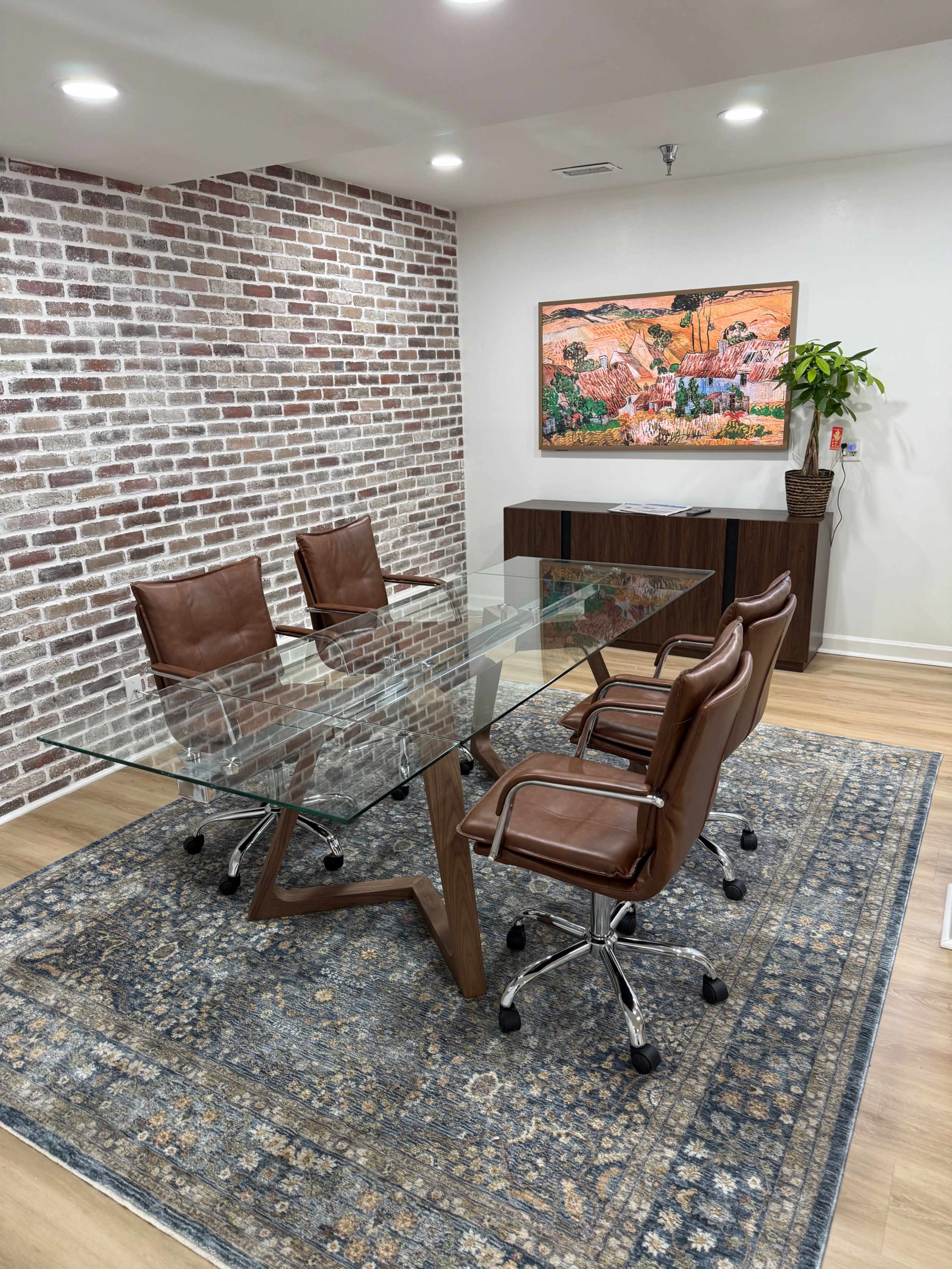 The image shows a modern conference room featuring a glass table surrounded by four brown leather chairs, a decorative wall with exposed brick, and a colorful piece of artwork on the wall.