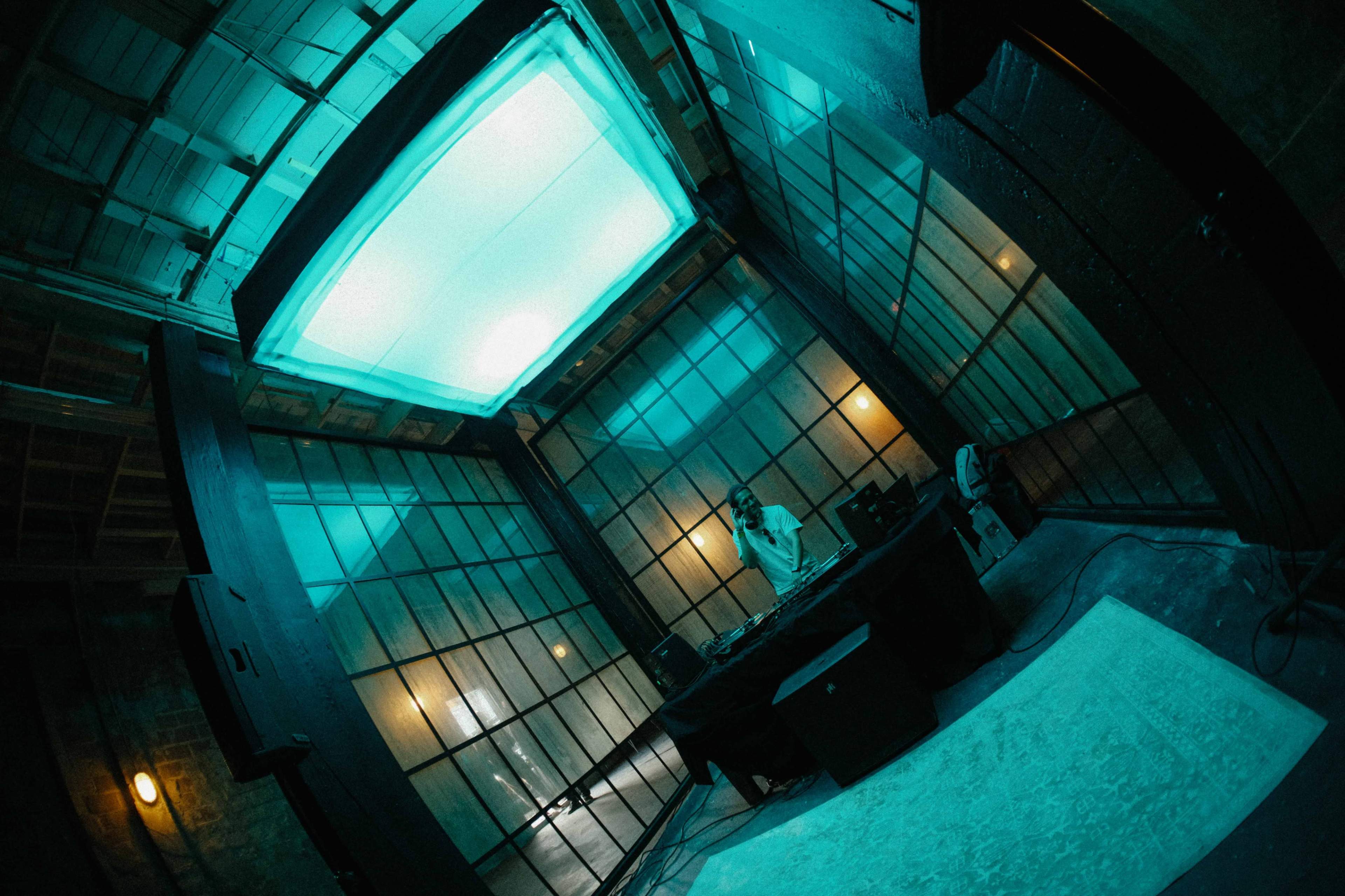 A DJ performs at a table in a dimly lit room with large glass windows and a glowing blue ceiling.