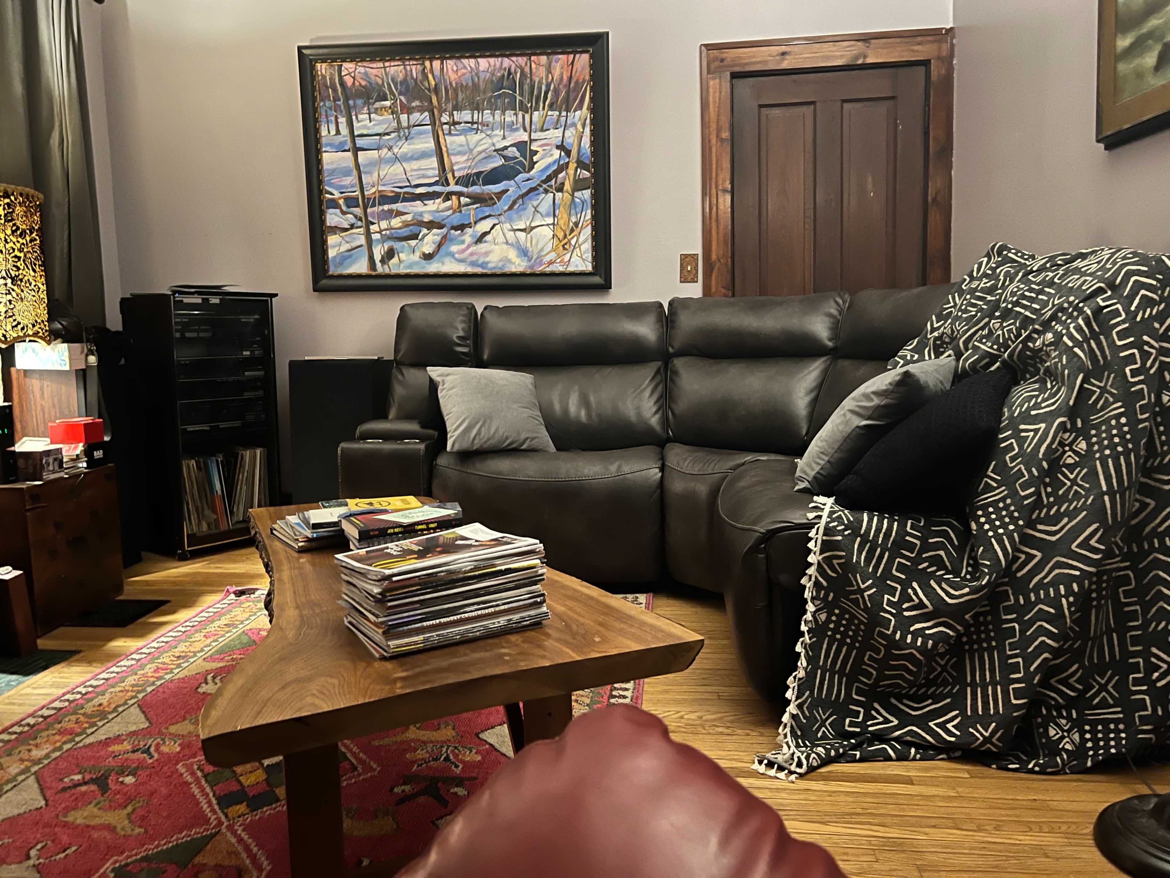 The cozy living room features a large sectional sofa, a wooden coffee table stacked with magazines, and a colorful area rug, with a painting of a winter landscape hanging on the wall.