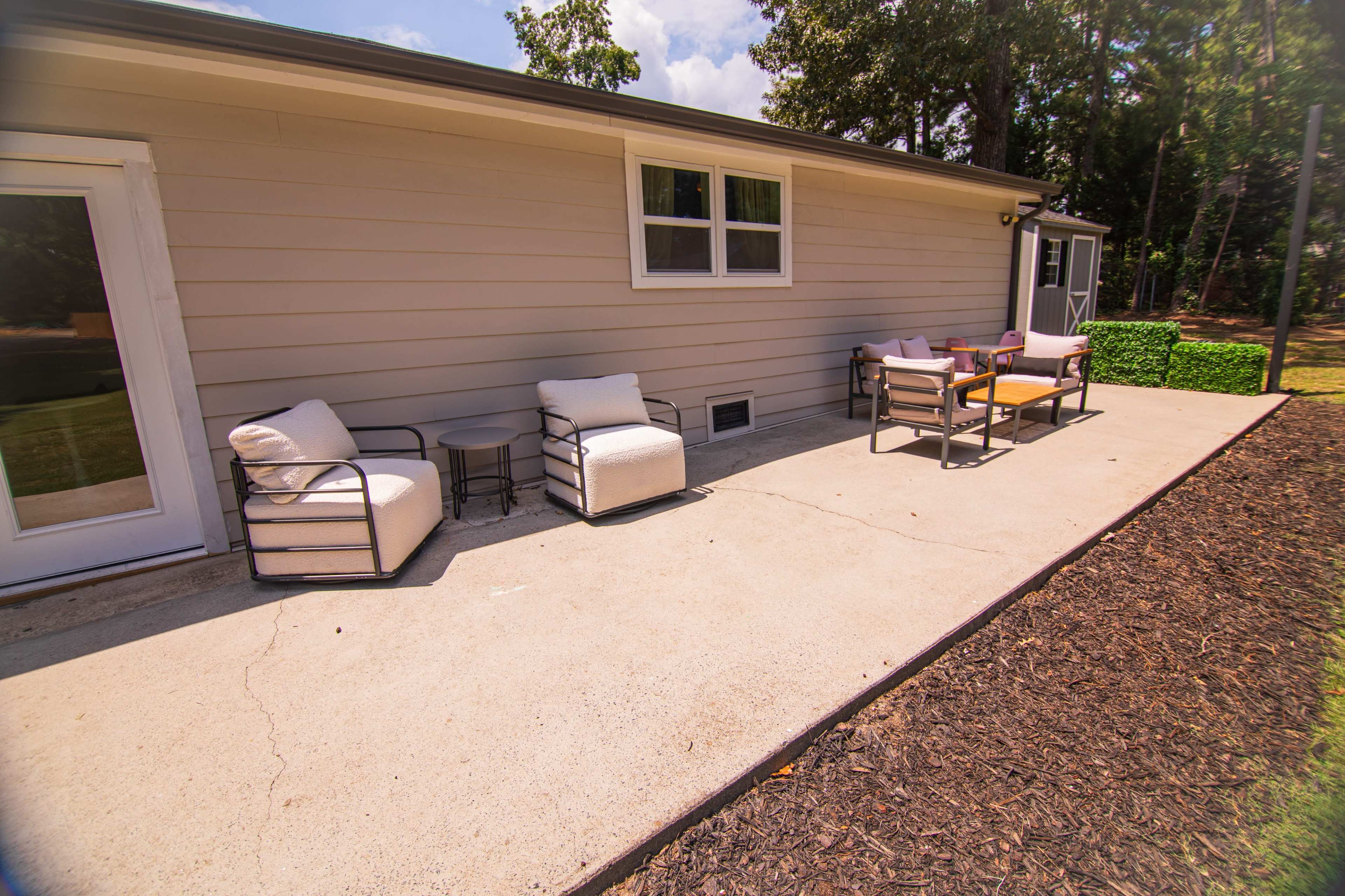 A patio features two chairs and a small table alongside a house, with landscaping elements nearby.