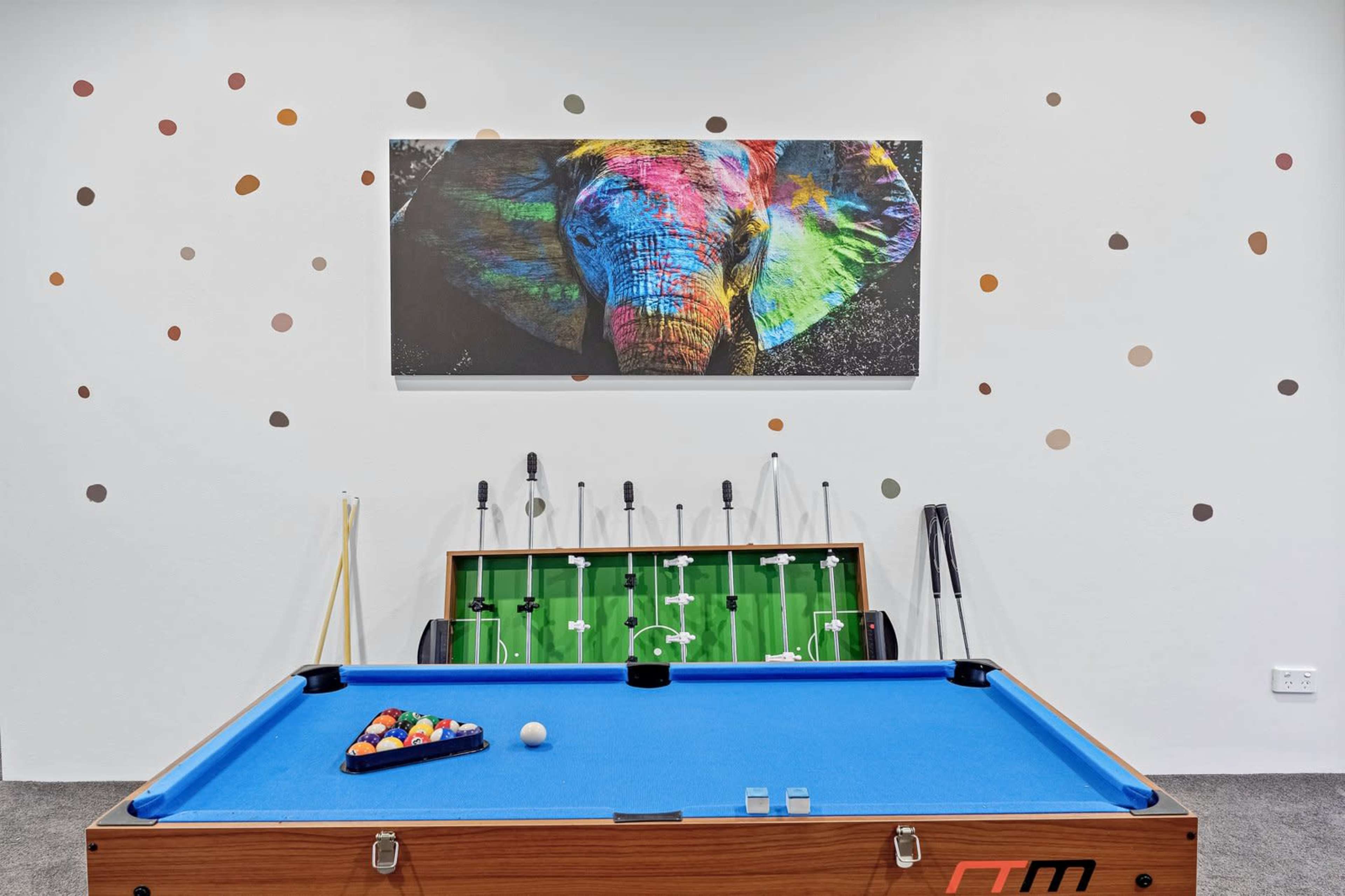The image shows a game room featuring a blue pool table, a foosball table, and a colorful elephant artwork on the wall.