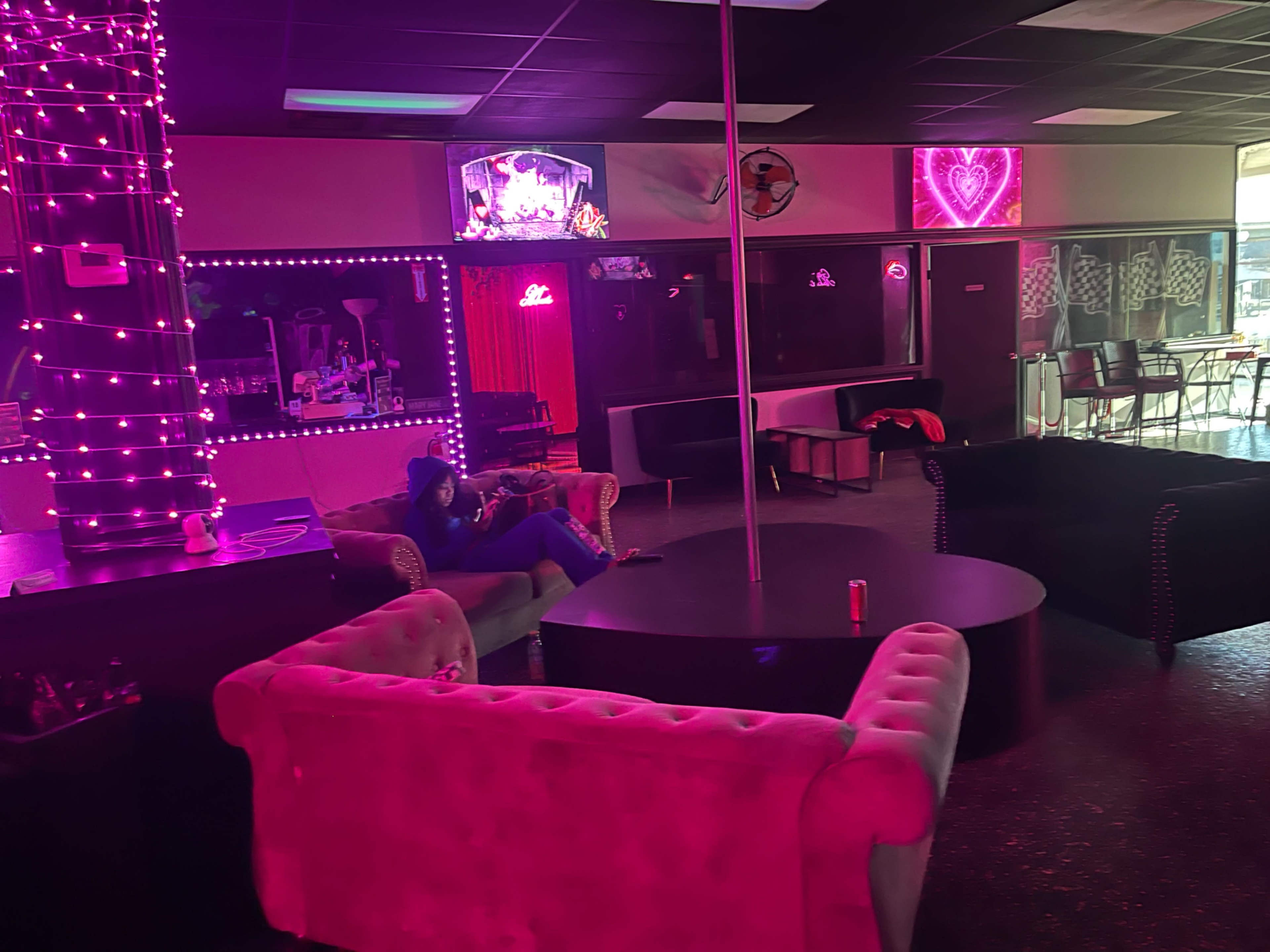 A dimly lit lounge area with pink neon lights, featuring couches and a central table, and a person sitting on a couch.