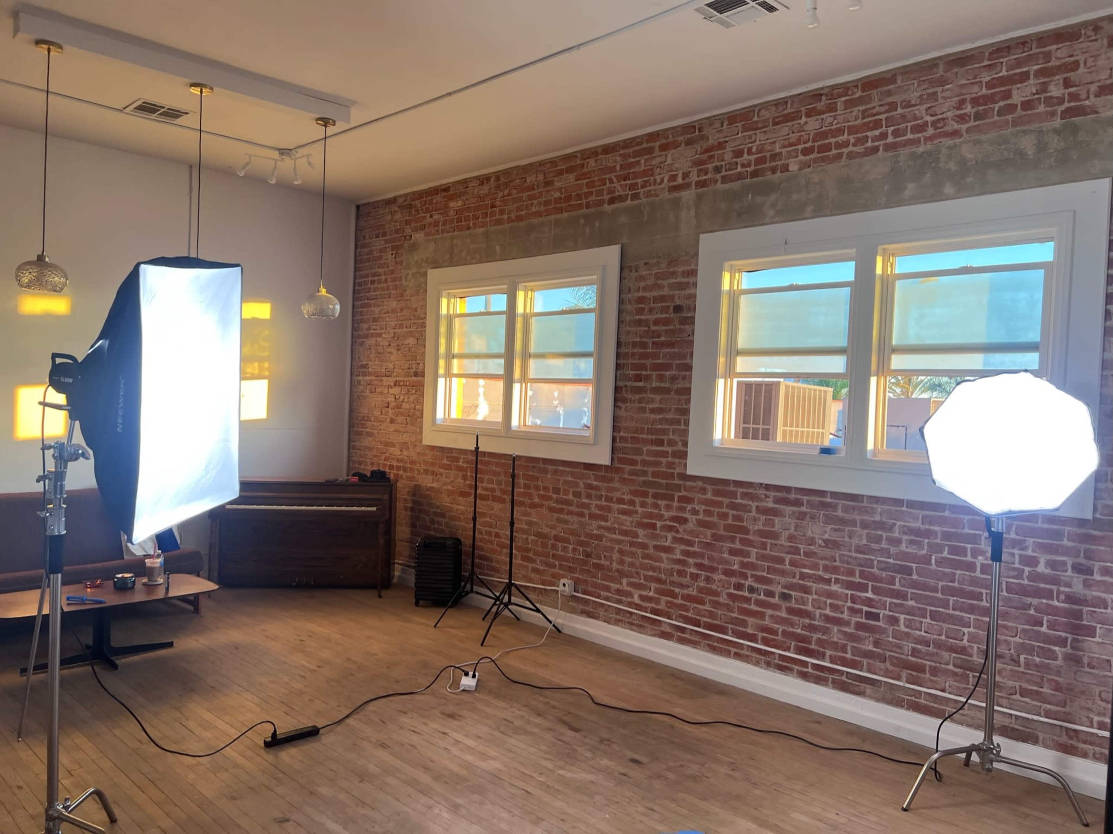 The image shows a well-lit room with exposed brick walls, featuring two lighting setups—one softbox and one umbrella light—alongside a wooden piano and scattered photography equipment.