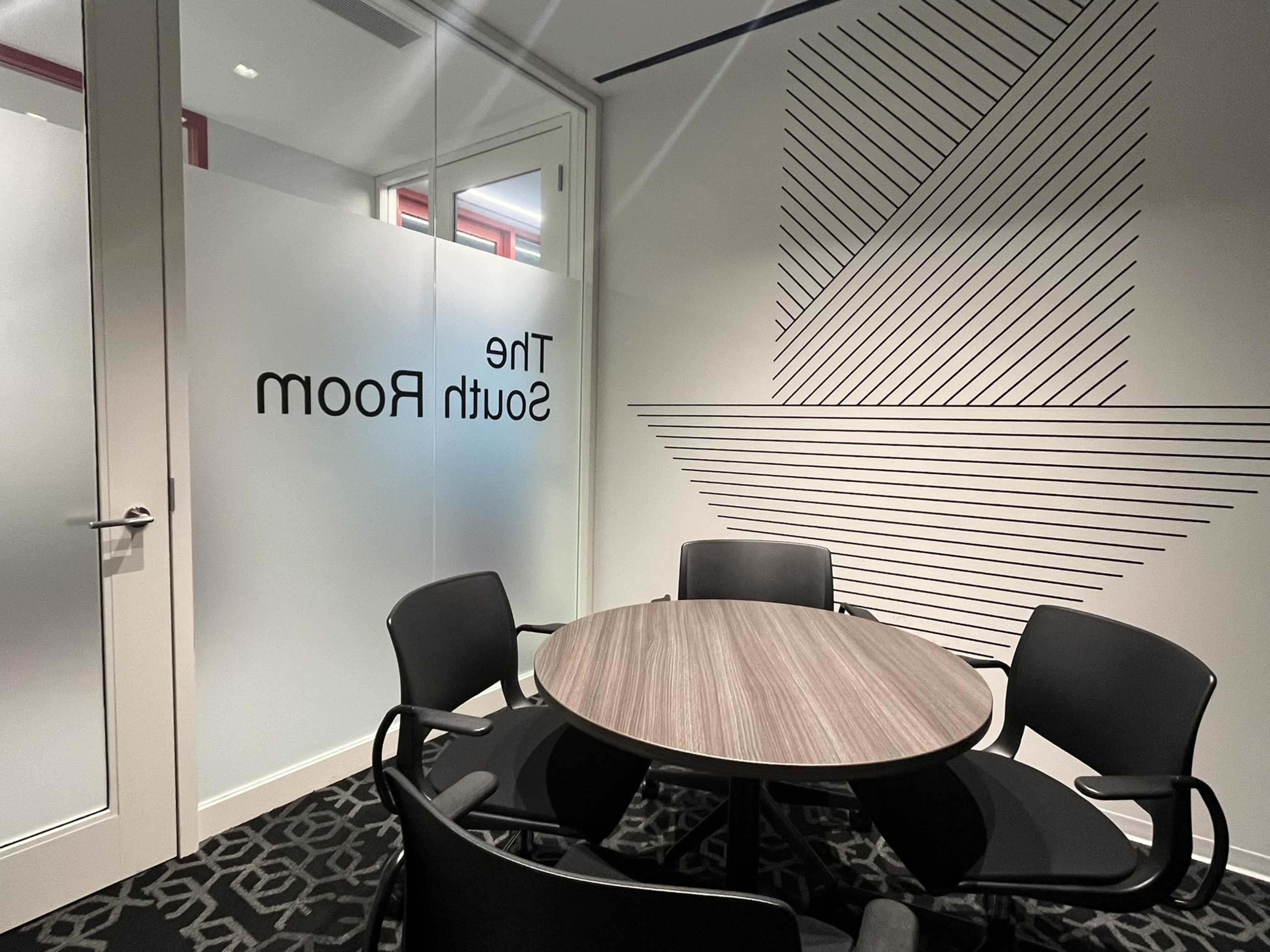 The image shows a modern meeting room with a round table surrounded by four black chairs, featuring a frosted glass door with the text "The South Room" and geometric lines on the wall.