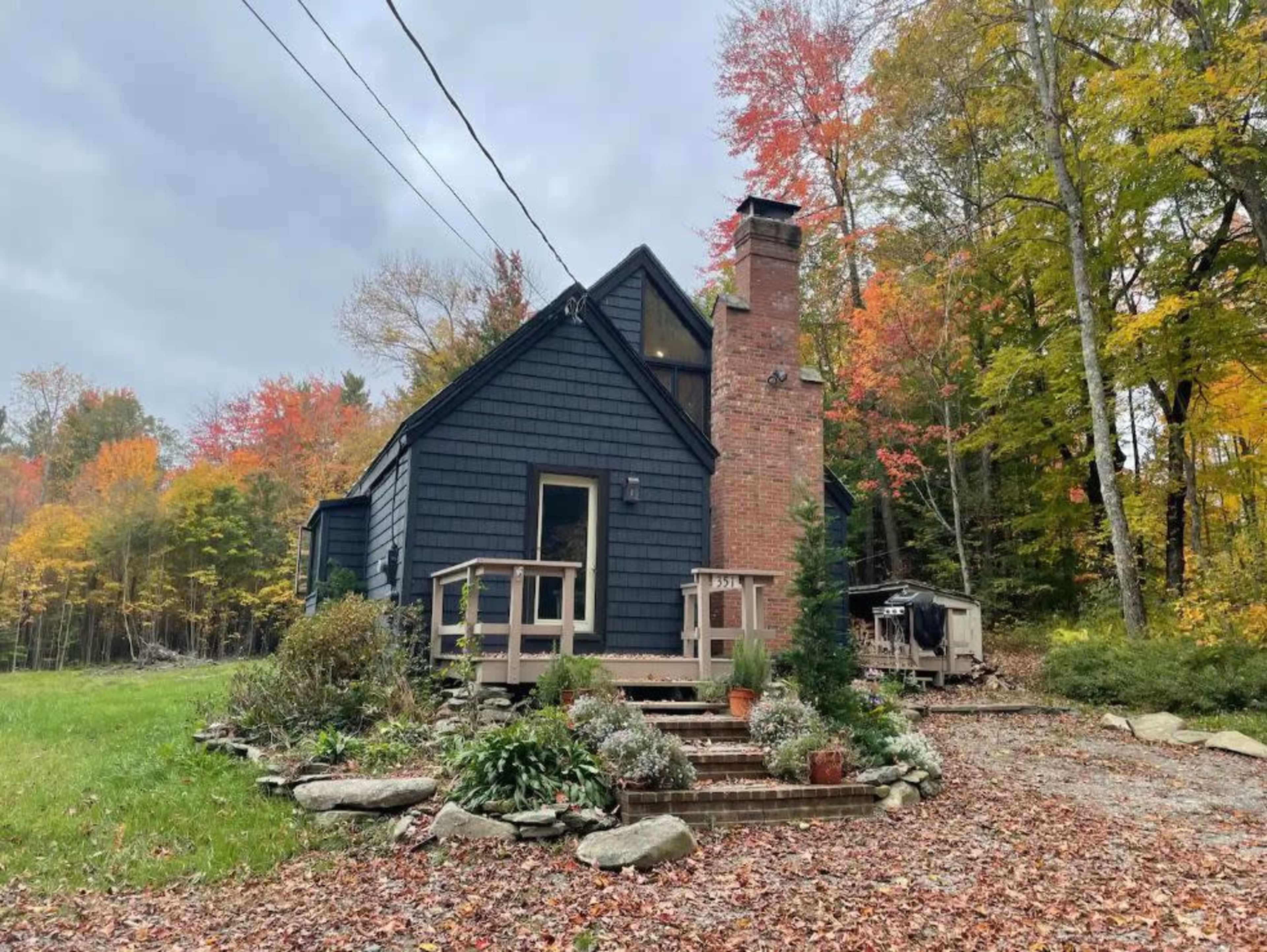 A two-story house with a brick chimney and dark siding is surrounded by trees with autumn foliage.