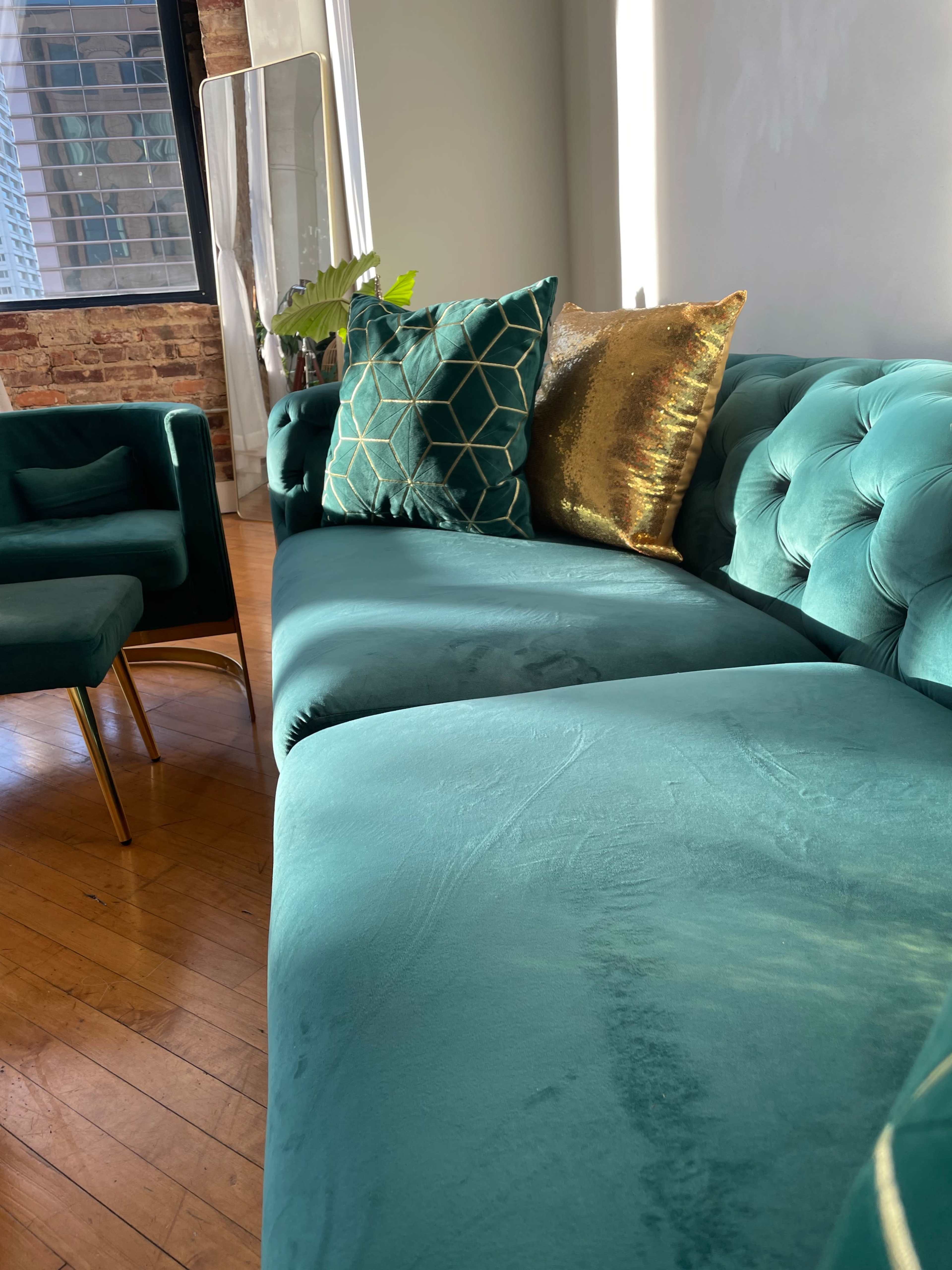 A tufted teal couch with decorative pillows sits in a well-lit room beside a large window and a tall mirror.