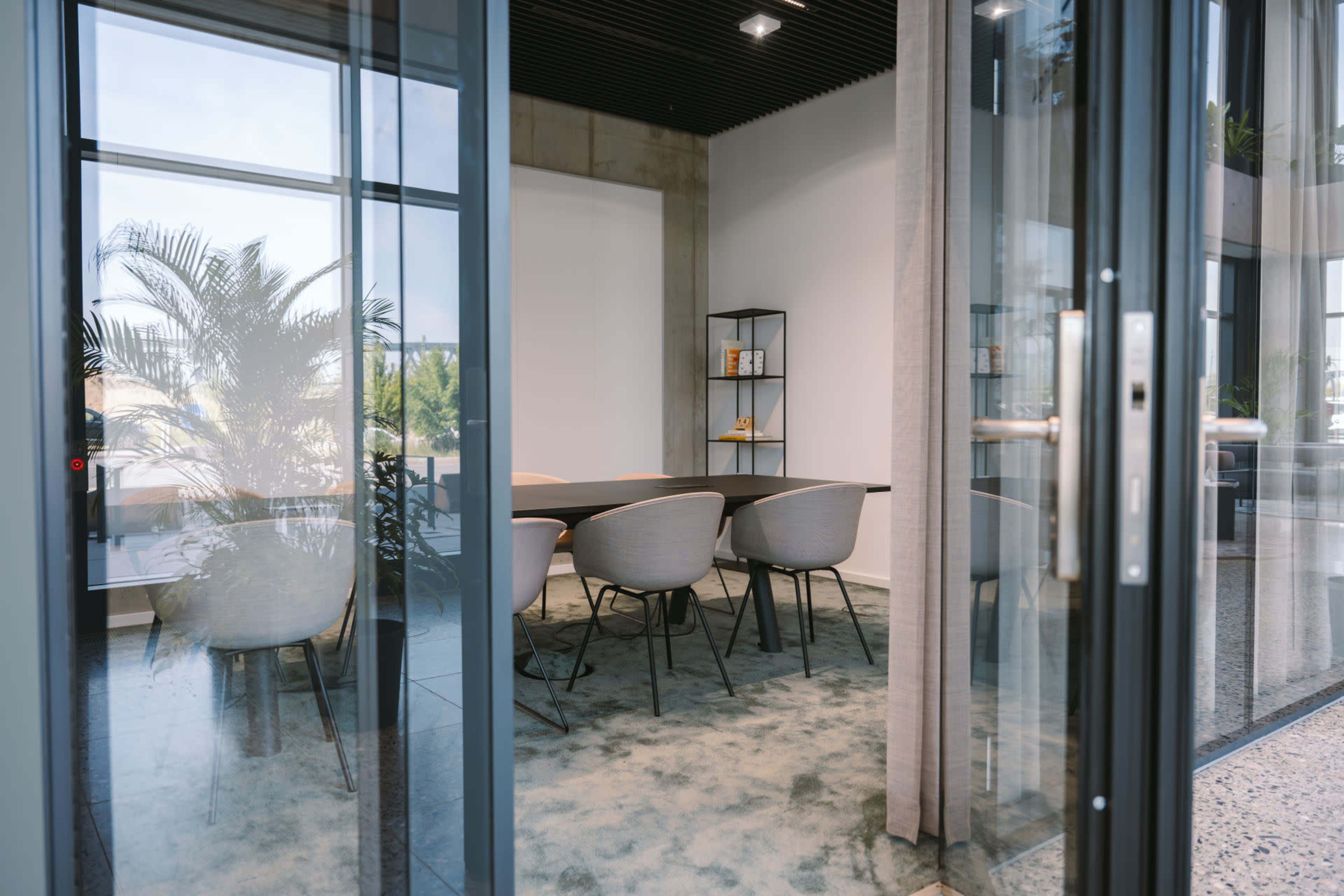 A glass door leads into a modern conference room featuring a large table and several chairs, with a bookshelf on one wall and greenery visible outside.