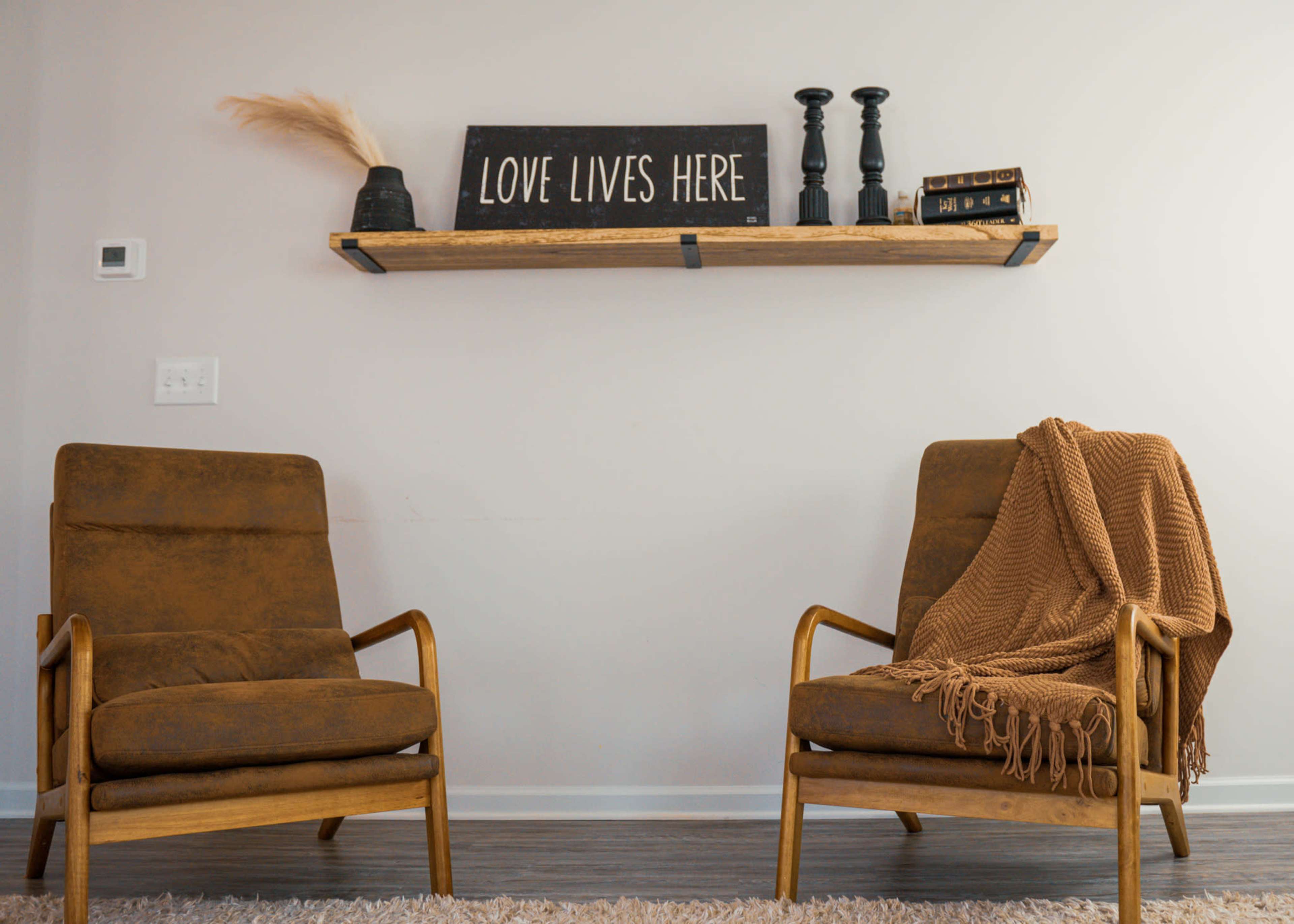 Two brown chairs are positioned facing each other beneath a wooden shelf adorned with a sign that reads "LOVE LIVES HERE," two candles, and a stack of books.