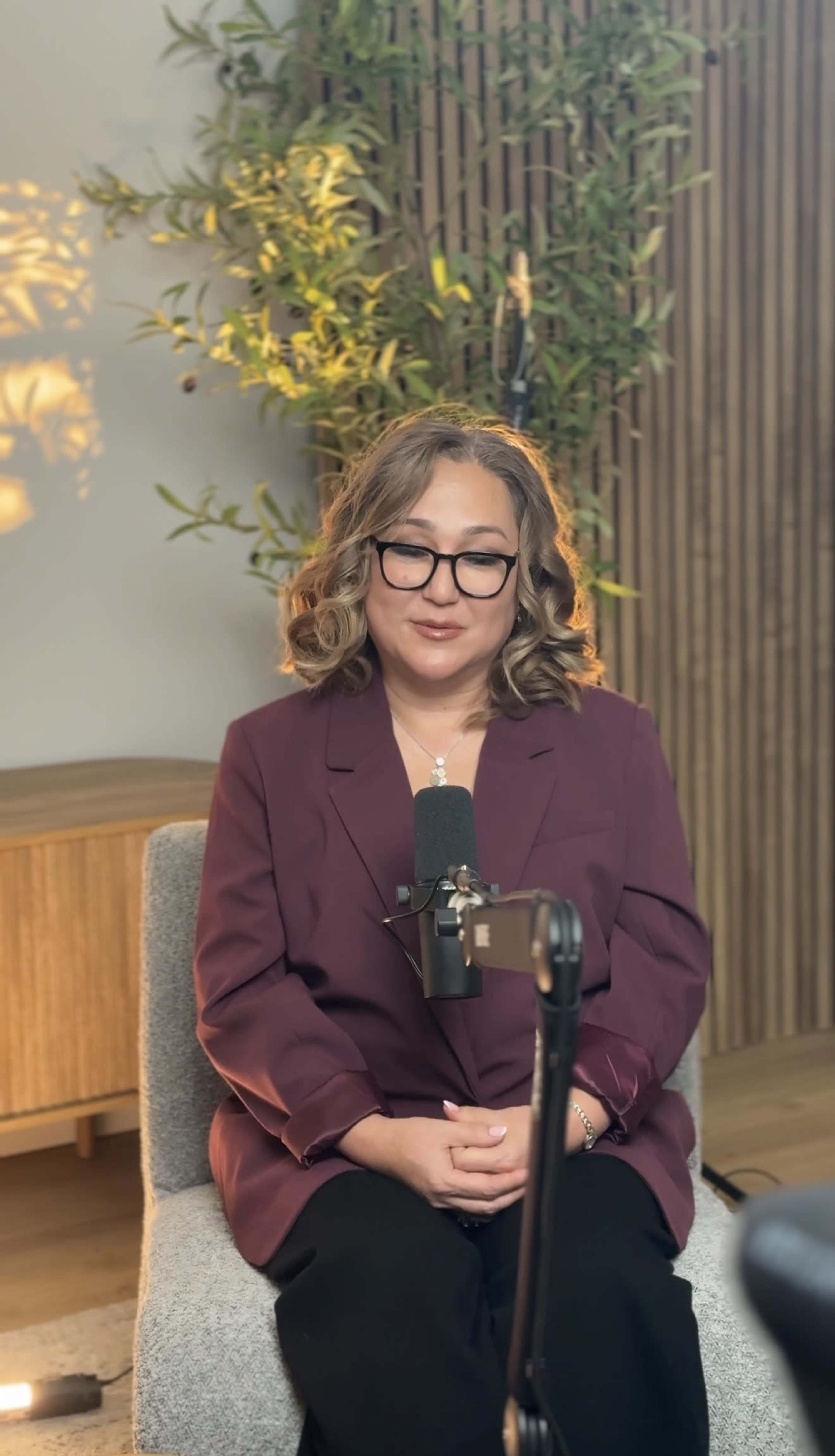 A woman with curly hair sits in a chair in front of a microphone, surrounded by greenery and wooden decor.