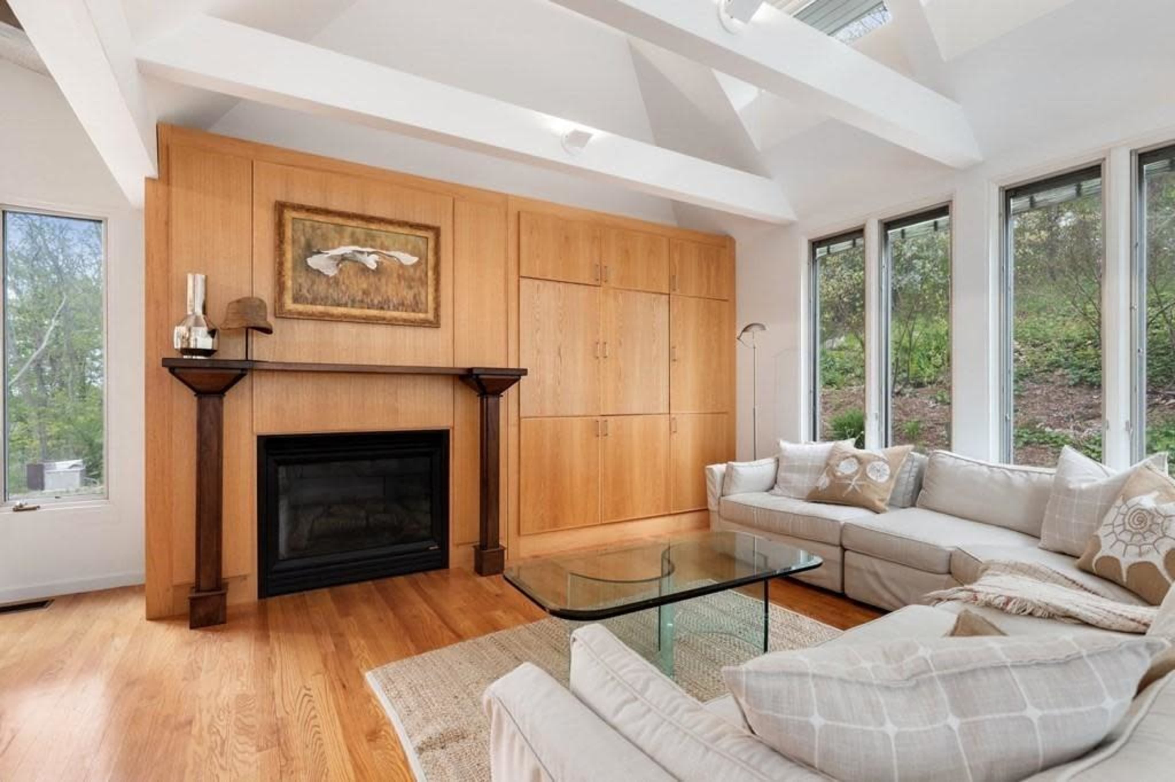 The room features a light-colored sectional sofa, a glass coffee table, and a wooden wall unit with a framed artwork above a black fireplace.