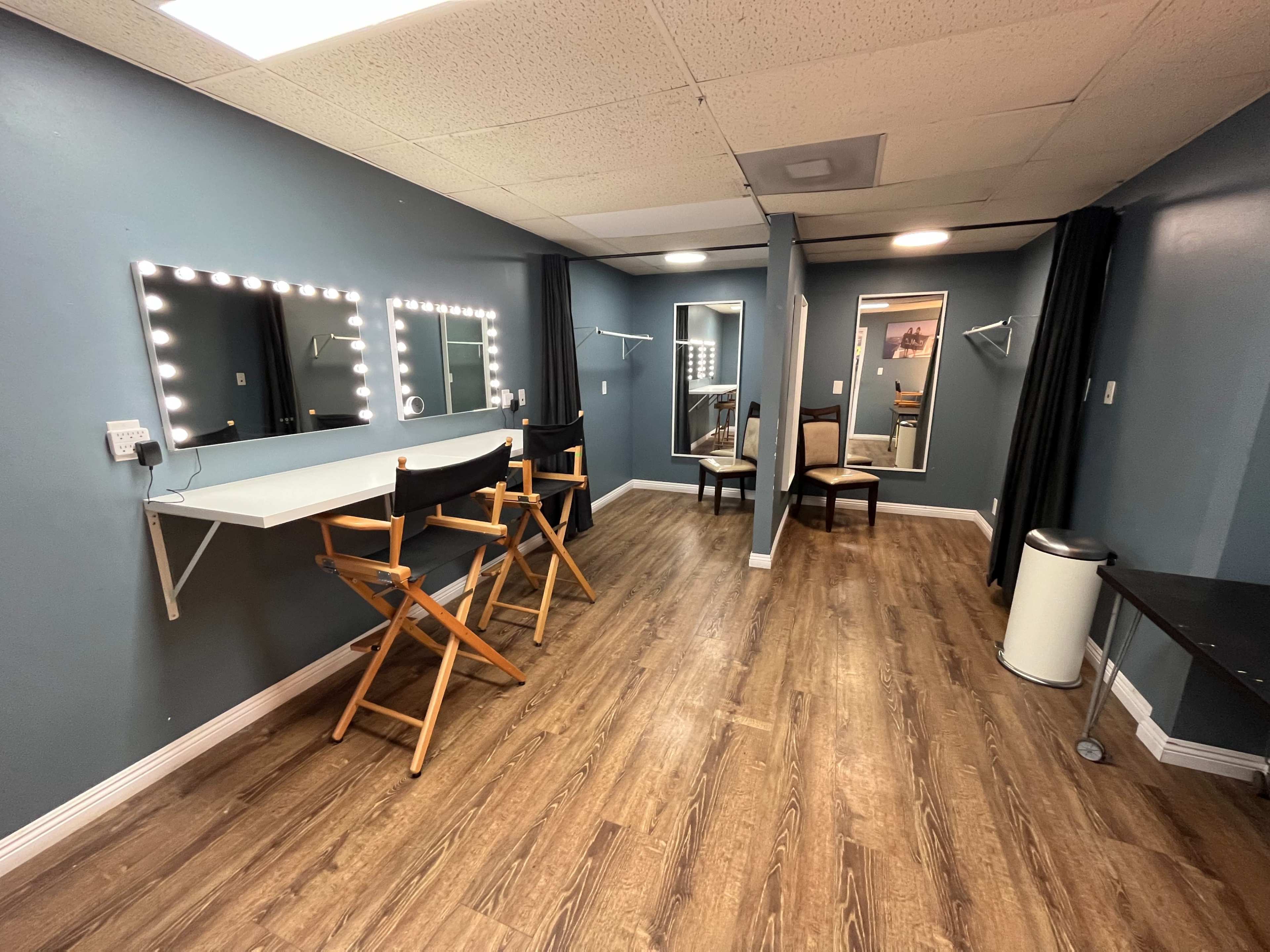 A well-lit dressing room with three makeup mirrors, two chairs, and a mirror at the far end.