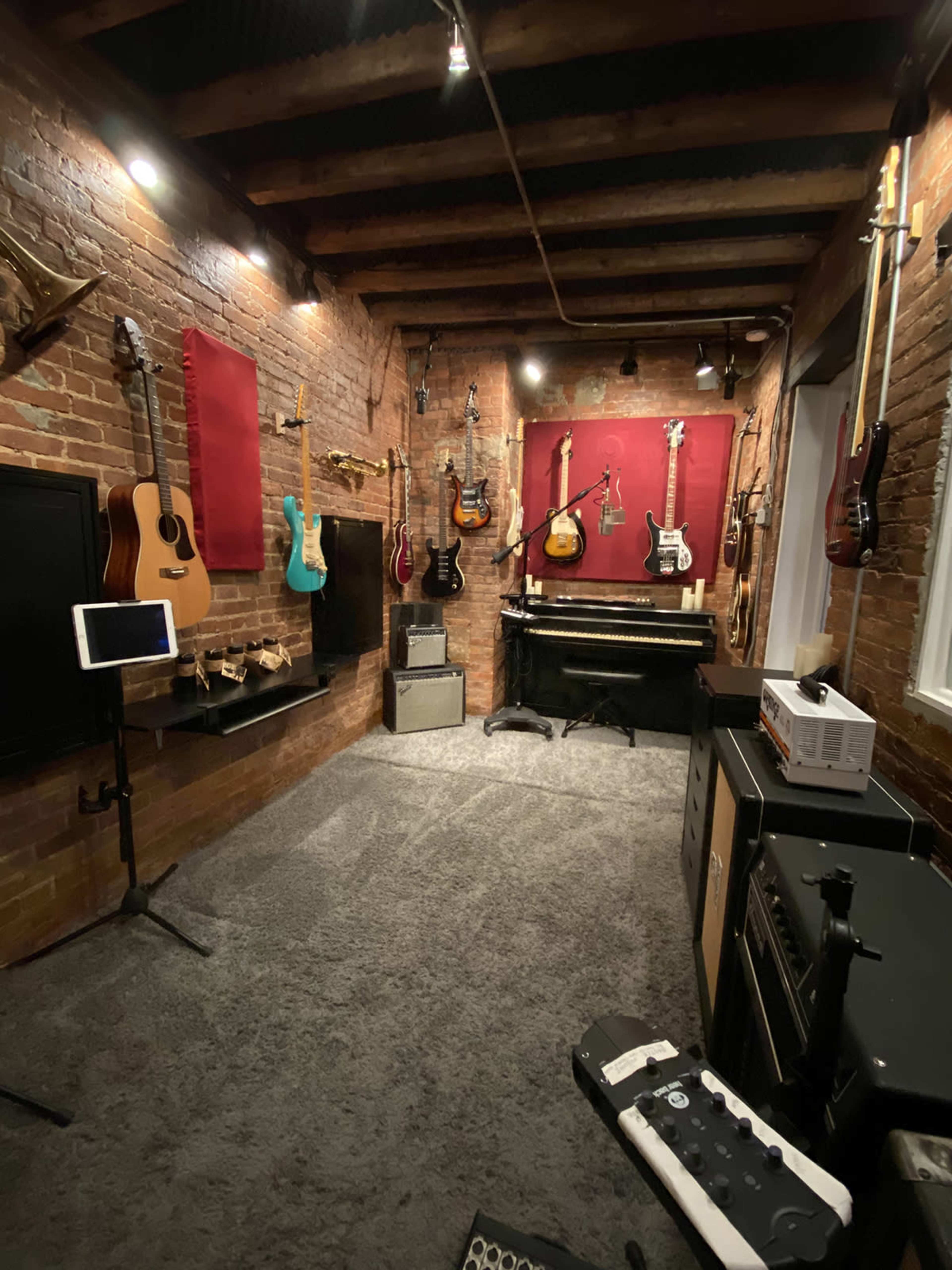A music studio featuring an assortment of guitars hanging on brick walls, a piano in the corner, and various amplifiers and equipment on the floor.
