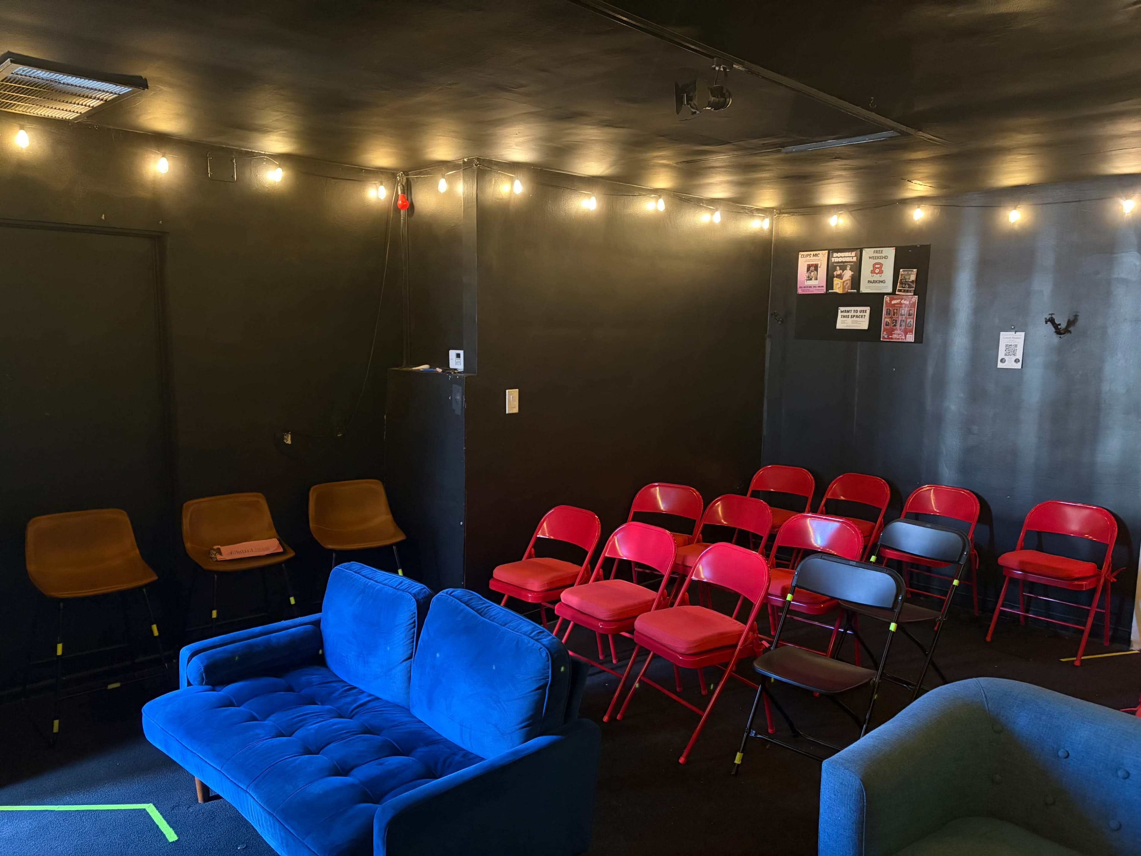 The image shows a small, dimly lit room with several rows of red folding chairs, a few brown chairs, and a blue couch, all surrounded by string lights on the walls.