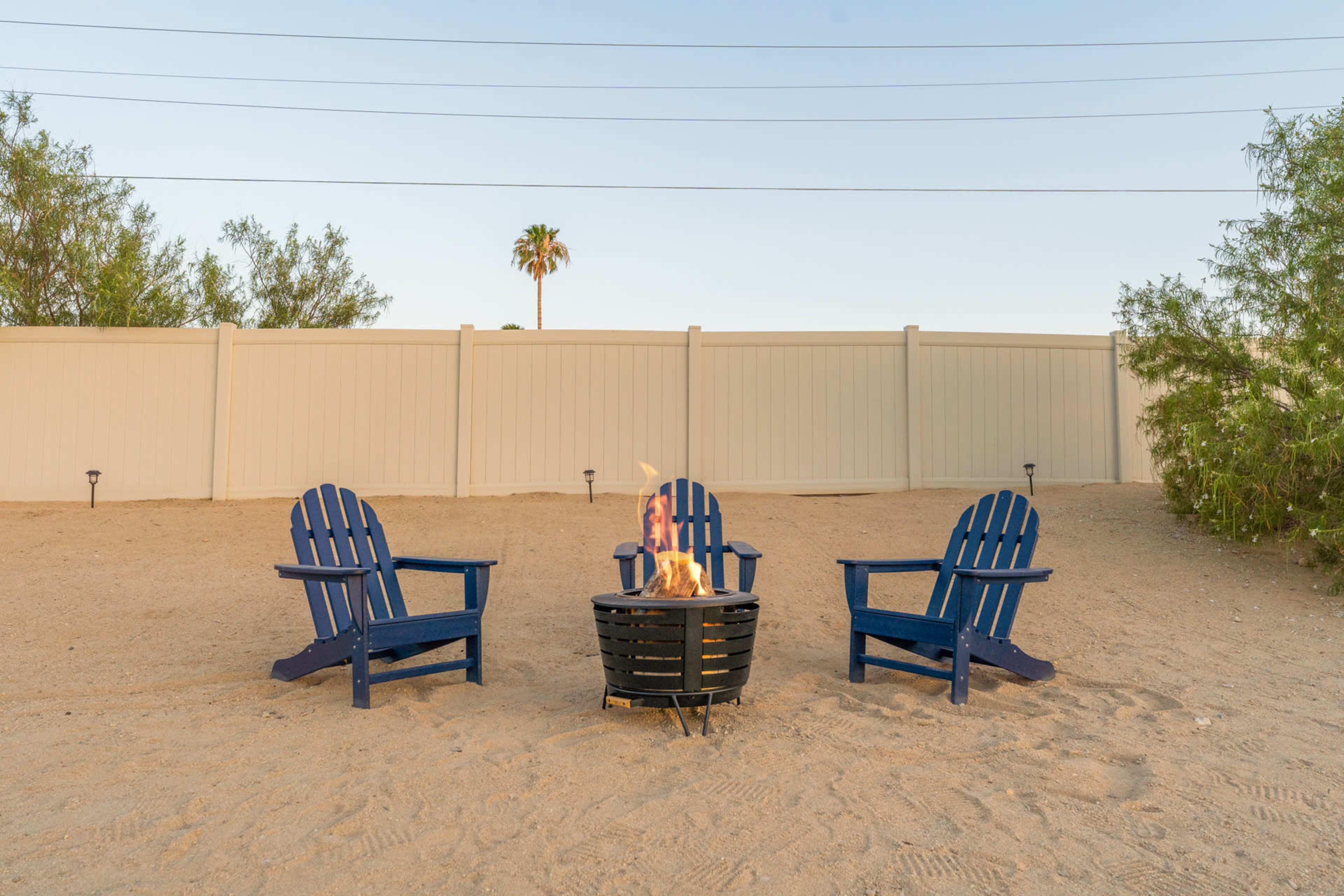 Three blue Adirondack chairs are arranged around a fire pit on a sandy area, with a palm tree visible in the background behind a white fence.