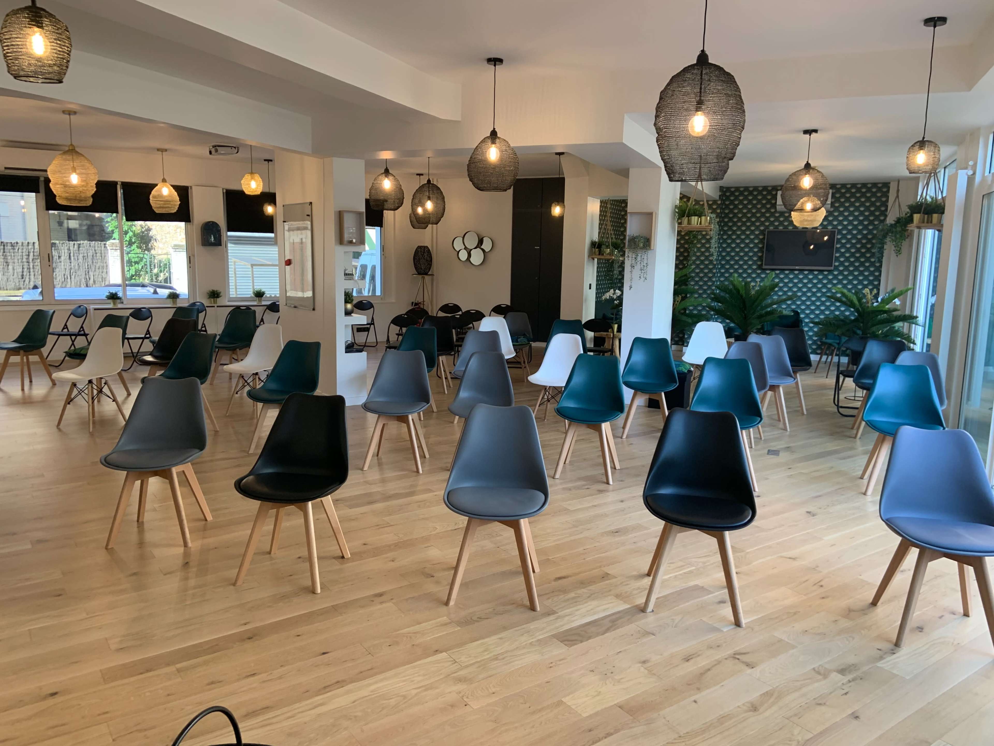 A spacious room with wooden flooring arranged with several colorful chairs featuring wooden legs, surrounded by decorative pendant lights and plants.