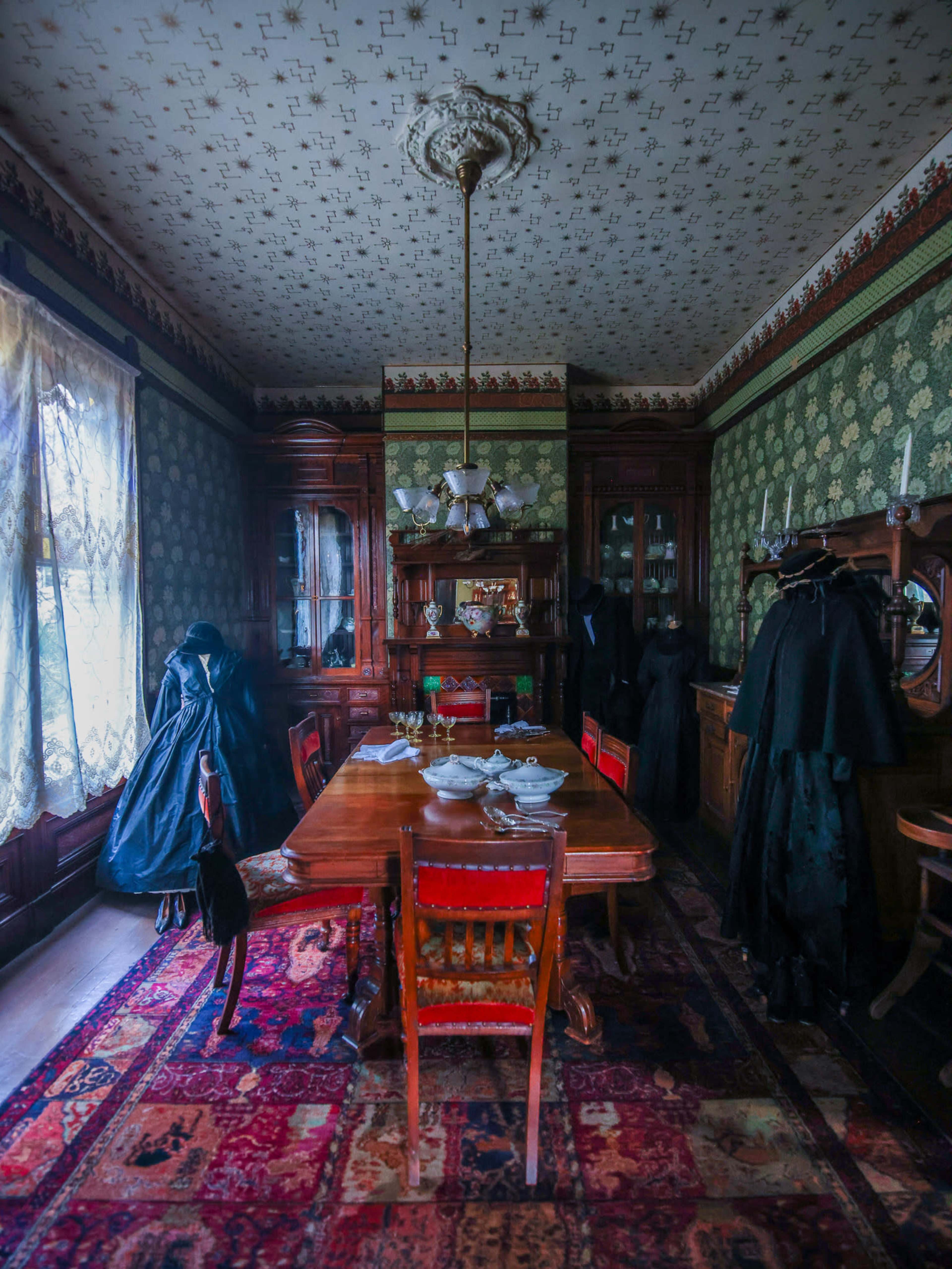 The image shows a dimly lit vintage dining room with a large wooden table surrounded by chairs, set against a backdrop of ornate wallpaper and a patterned rug.