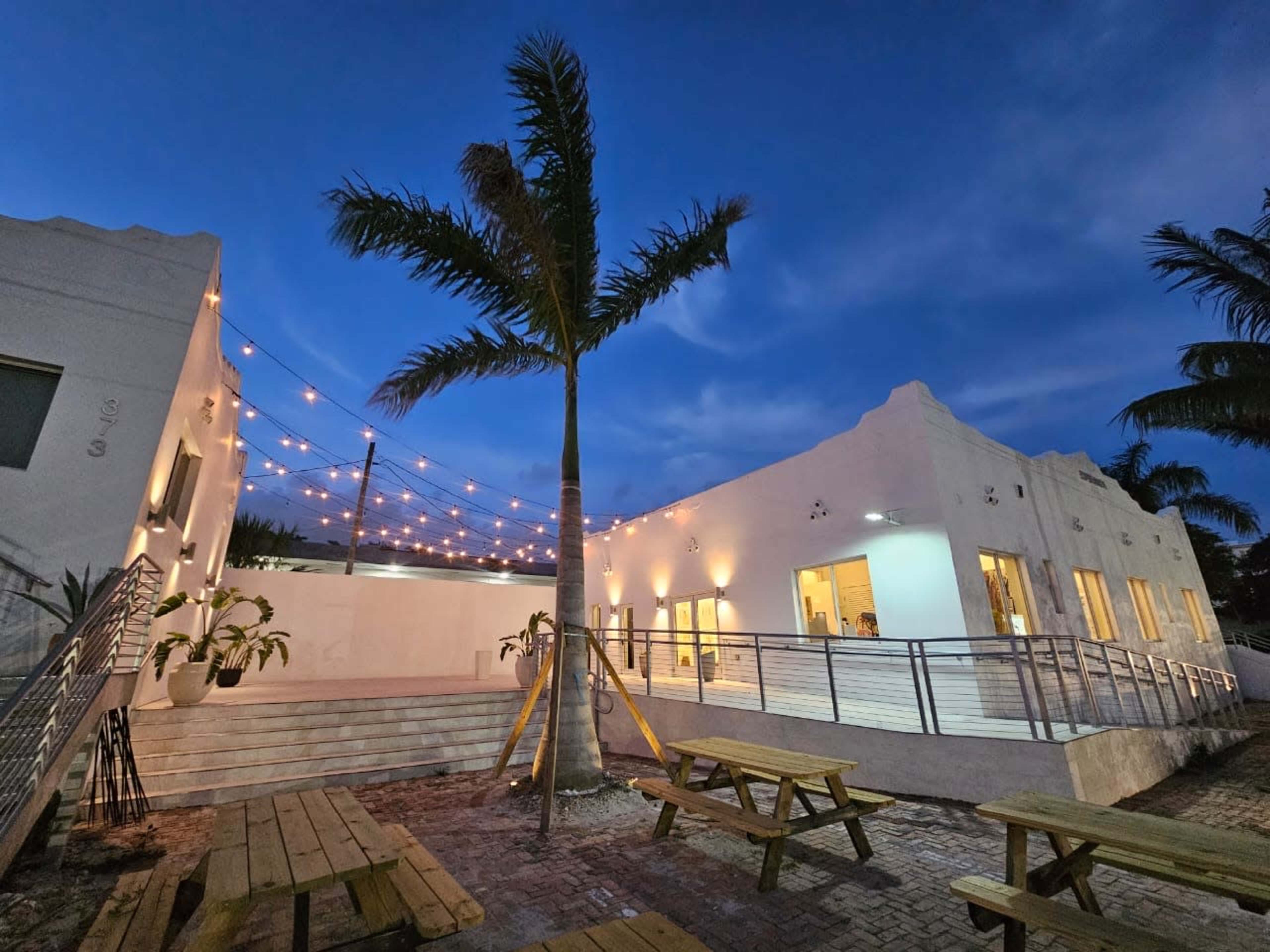 A modern building with large windows is illuminated by string lights in an outdoor seating area surrounded by palm trees at dusk.