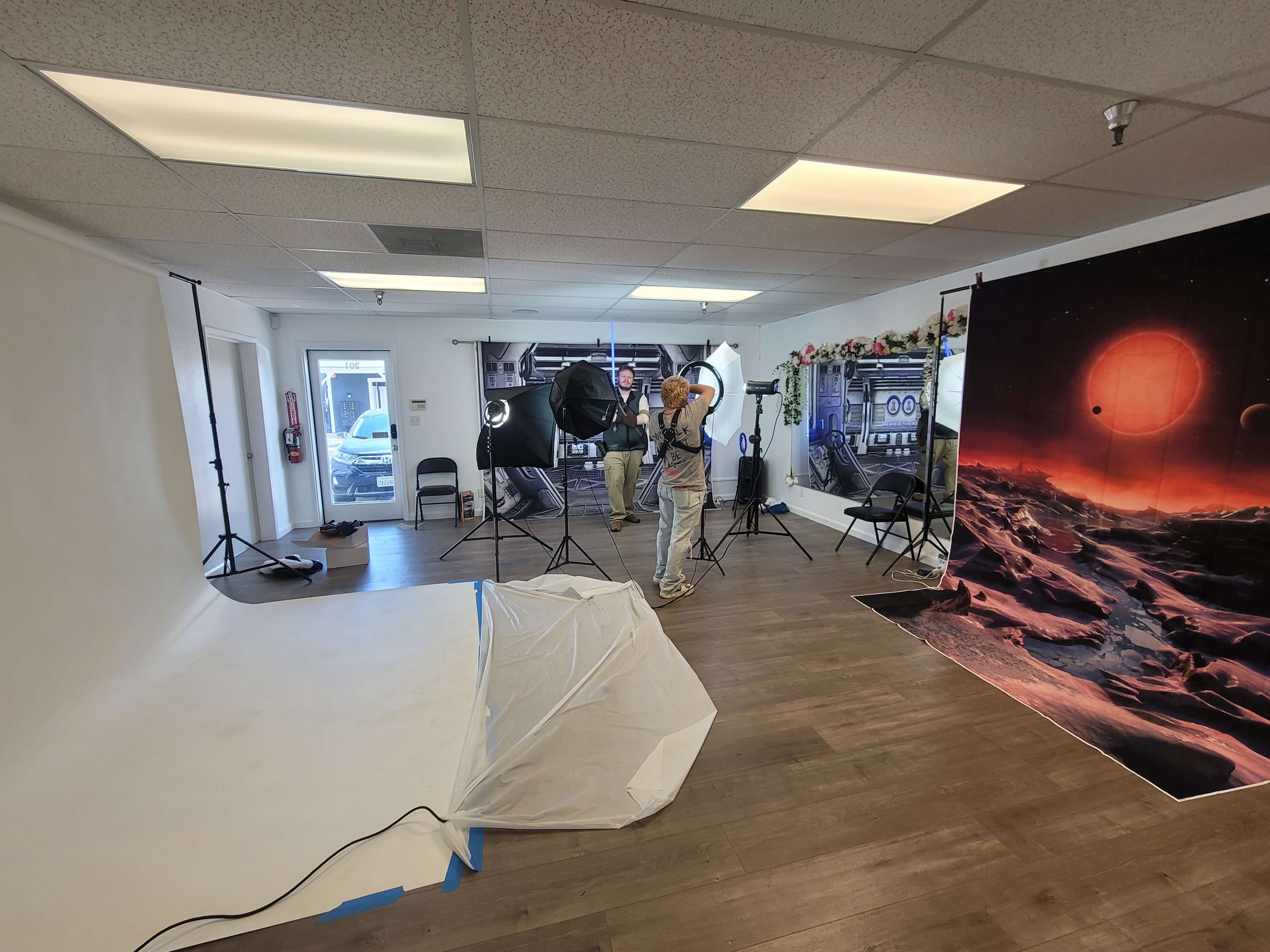 A photographer is setting up a lighting kit in a studio with a backdrop of a space scene and a blank white wall.