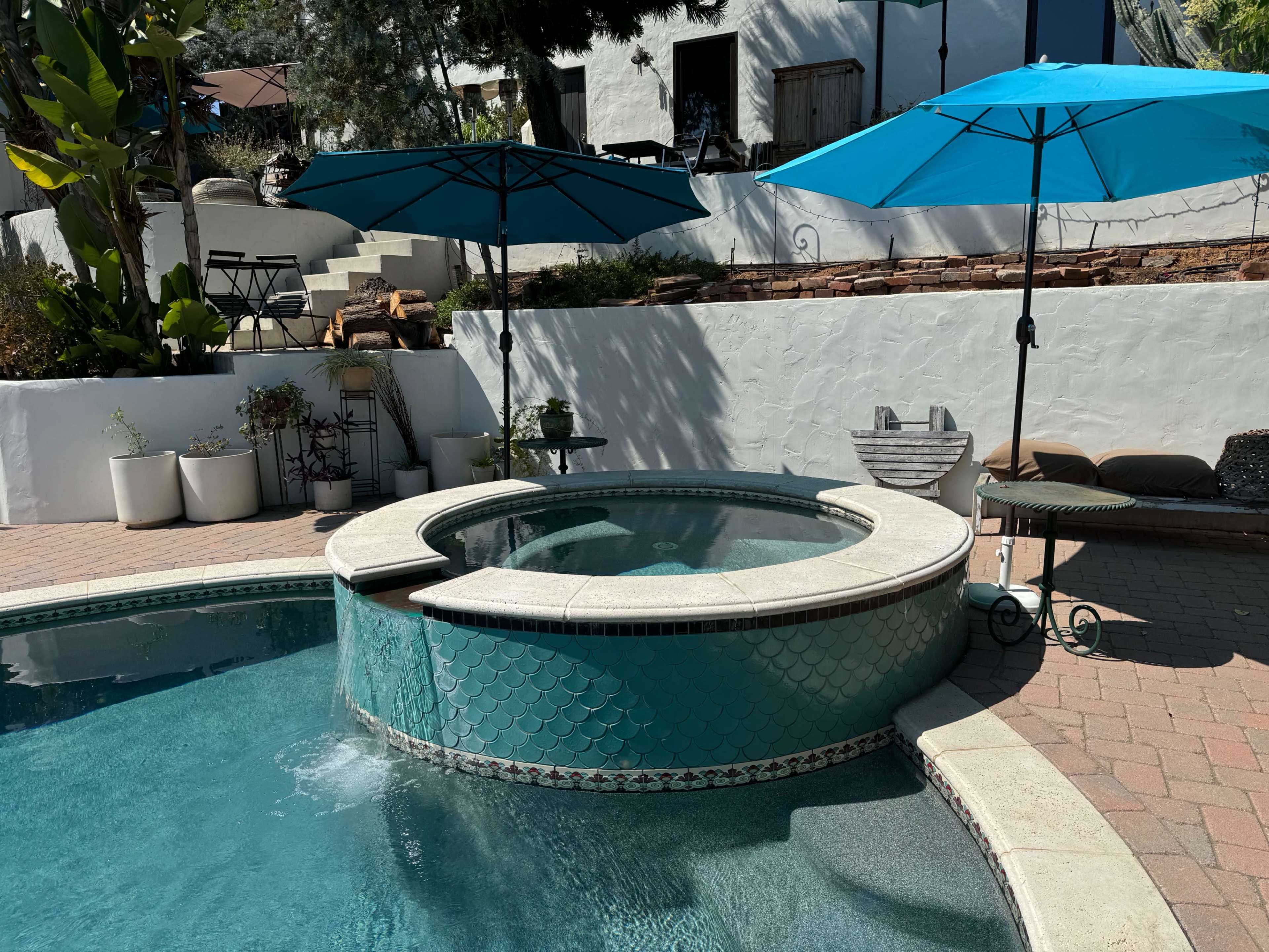 The image shows a spa area adjacent to a swimming pool, featuring two large turquoise umbrellas and surrounded by potted plants.